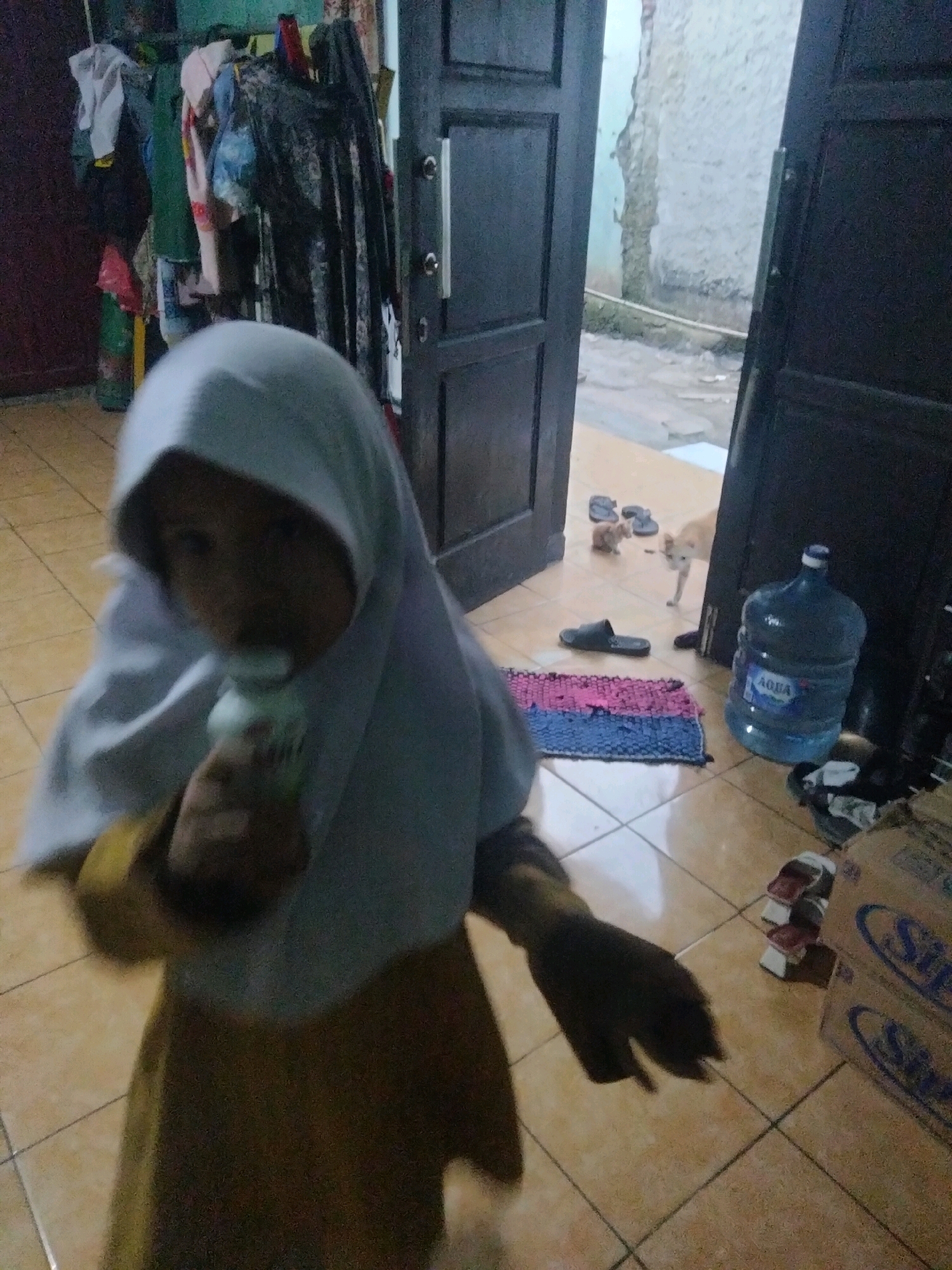 In Suka Sari, Indonesia, a young girl wearing a light blue hijab and a yellow dress is holding a white bottle near her mouth. She is standing in a tiled room with a dark brown door open to an outdoor area. In the outdoor area, two cats are present: one is light brown and the other is orange and white. A large blue water jug with the label "AQUA" is visible to the right of the door. Several pairs of black sandals are scattered on the floor near the doorway. There are also stacks of cardboard boxes with the blue "SUPER" logo, one of which has "SP" visible. Clothing is hanging on a rack in the background, and a small pink and blue rug is on the floor. The walls are a light, textured color, and the ground outside appears to be dirt.