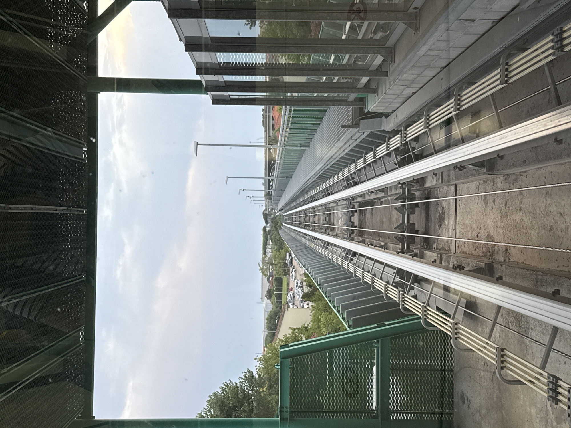 This image captures a view from a modern train or monorail, likely operating in the city of Pisa, Italy, as indicated by external context. The perspective is looking forward along a set of elevated train tracks made of metal, running parallel to each other. The tracks are situated on a raised platform with a concrete base and a grey metal walkway alongside. To the right, a contemporary building with large glass panels and a metal frame is visible, reflecting the sky and surrounding structures. On the left side of the frame, a green metal barrier with a mesh-like texture is present, suggesting a safety feature for the elevated track. Beyond the tracks and to the left, a parking lot filled with various vehicles is discernible, with trees and some low-rise buildings in the background. The sky is overcast with a soft, diffused light, indicating a cloudy day. The overall scene suggests an urban or suburban environment with public transportation infrastructure.