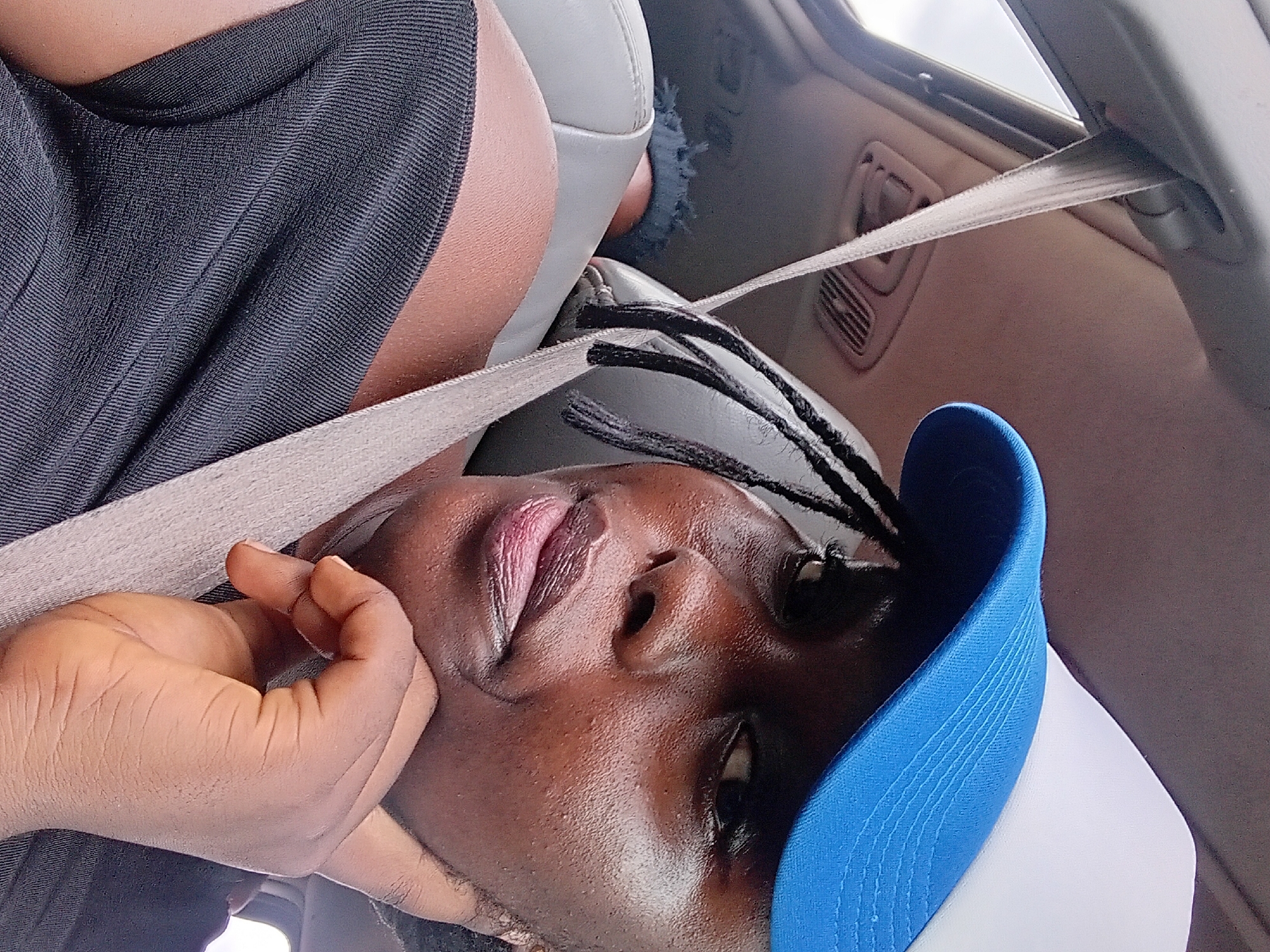 A close-up, waist-up shot captures a young Black woman in the passenger seat of a car, wearing a black off-the-shoulder top and a blue and white baseball cap. She rests her chin on her hand, her gaze directed slightly to the right of the camera. Her braided hair is visible beneath the cap. The seatbelt is across her chest. The car interior, including the headliner and part of the backseat, is visible. The background suggests an outdoor environment, possibly Adoma, Nigeria, though this is not definitively confirmed by visual cues.