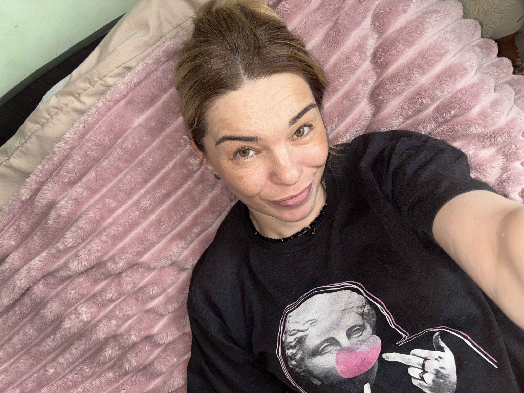 A woman is captured in a close-up selfie, lying down indoors in Vsevolozhsk, Russia. She appears to be in her 30s or 40s, with light brown hair styled in a bun, and light brown or hazel eyes. She sports a subtle, content smile. She is wearing a black long-sleeved top adorned with a distinctive graphic print. The graphic features a grayscale rendition of a classical bust, resembling ancient Roman or Greek sculpture, wearing a vibrant pink surgical mask. A hand is depicted in the graphic, pointing towards the masked bust with its index finger extended. She also wears a dark, possibly beaded, necklace. The woman is lying on a soft, plush, pink blanket or comforter that has a rippled texture. To her left, a light beige sheet or pillowcase is visible alongside a dark furniture edge, with a pale green wall section in the upper left corner of the frame. The image conveys a casual and relaxed indoor moment.