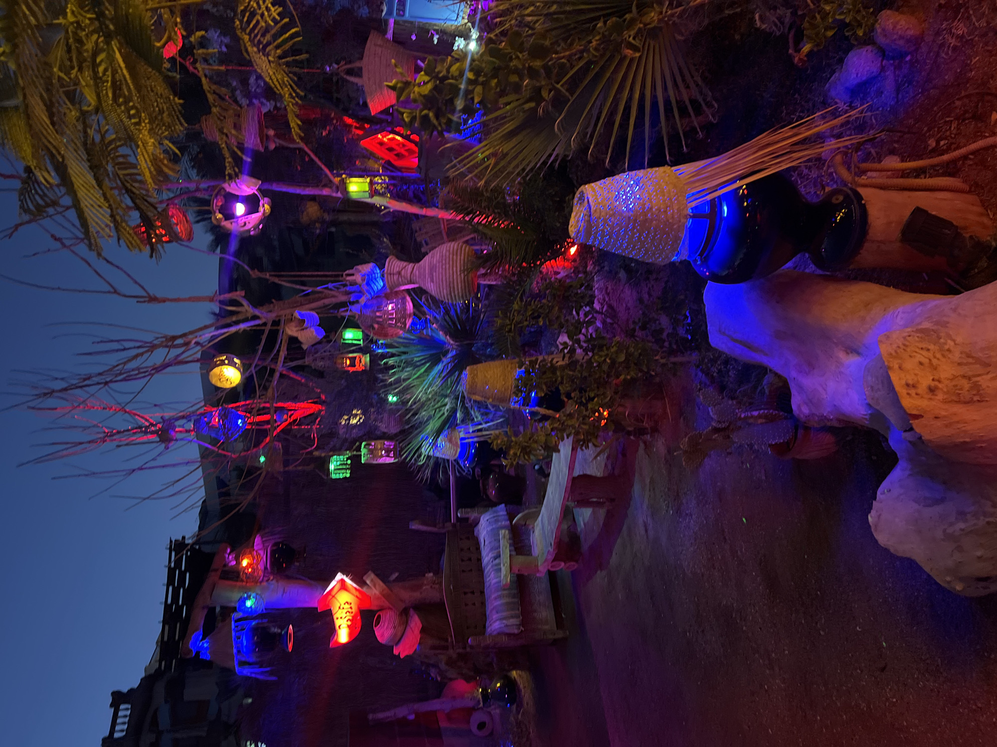 The image depicts a decorative outdoor setting at dusk or night, illuminated by numerous colorful lights. The scene is richly textured with a variety of objects, suggesting a bohemian or artistic ambiance. In the foreground, to the right, a large dark vase is topped with a woven, conical shade that glows with blue light from within. This vase sits on a wooden stump, and a coiled hose is visible near its base. To the left of the vase, a large, pale, rough-hewn rock formation is illuminated by red and purple lights.
Further into the scene, a rustic wooden bench with a patterned cushion is positioned near a larger, more ornate wooden bench with carved details. Behind these benches, a tree with bare branches is adorned with an array of hanging lanterns and decorative objects. These include brightly colored glass lanterns in red, green, blue, and yellow, as well as woven baskets and other textured, possibly ceramic, items. Some of the lanterns are shaped like fish or have ornate cutouts. In the mid-ground, lush green foliage and spiky plants are visible, interspersed with more lights. The ground appears to be made of dark gravel or dirt.
In the background, a building structure with horizontal lines and a darker facade is visible against the twilight sky. The overall impression is one of an eclectic and vibrant outdoor display, likely intended for evening enjoyment.
This specific scene is located in the Naama Bay area of Sharm El Sheikh, Egypt, a popular tourist hub known for its nightlife, restaurants, and decorative streetscapes.