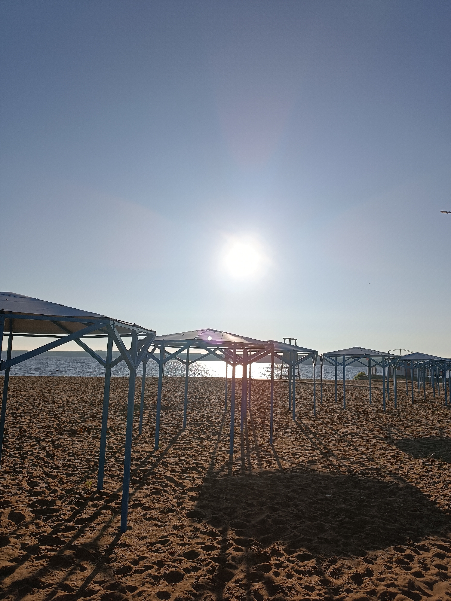 An outdoor, low-angle, eye-level shot captures a beach in Chaikovsky, Russia, on a clear day with the sun shining brightly. The foreground is dominated by a sandy beach with visible footprints and some sparse green vegetation. A series of empty, blue-framed canopies with light blue roofs are positioned in a line across the sand. These structures appear to be beach shelters. In the background, a body of water, likely a river or lake, stretches across the horizon, reflecting the sunlight. The sky is a clear, bright blue with a large, radiant sun positioned slightly to the left of the center. Shadows of the canopies are cast long across the sand, indicating the time of day. There are no people visible in the frame.