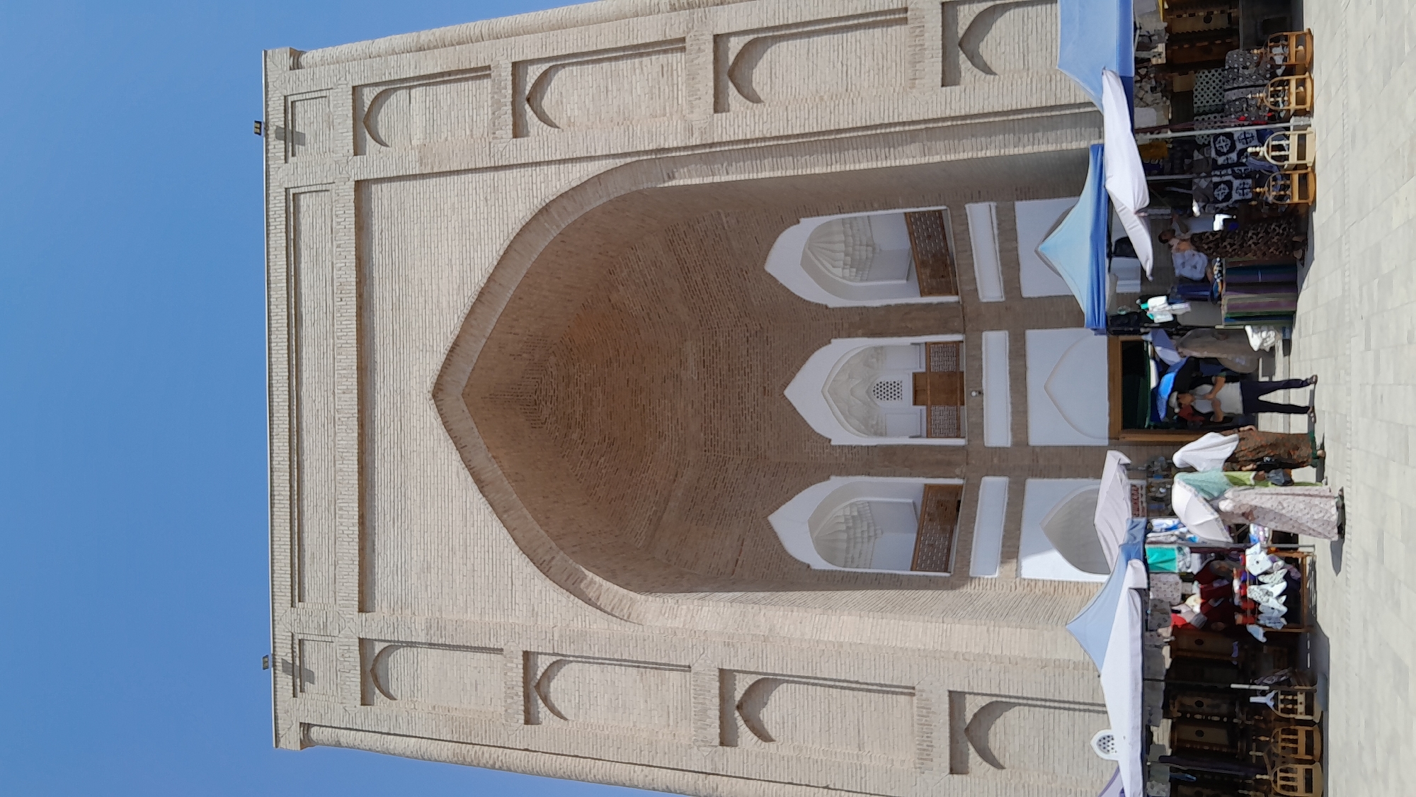 In Denov, Uzbekistan, a large, imposing brick building with an arched entrance dominates the frame. The architecture features multiple recessed arches and geometric patterns in the brickwork. In front of the building, a bustling marketplace is set up with stalls covered by colorful awnings. Several people are present, some browsing goods displayed on tables and racks, while others are walking or standing. The marketplace appears to be selling textiles, clothing, and decorative items. The sky is a clear, bright blue.