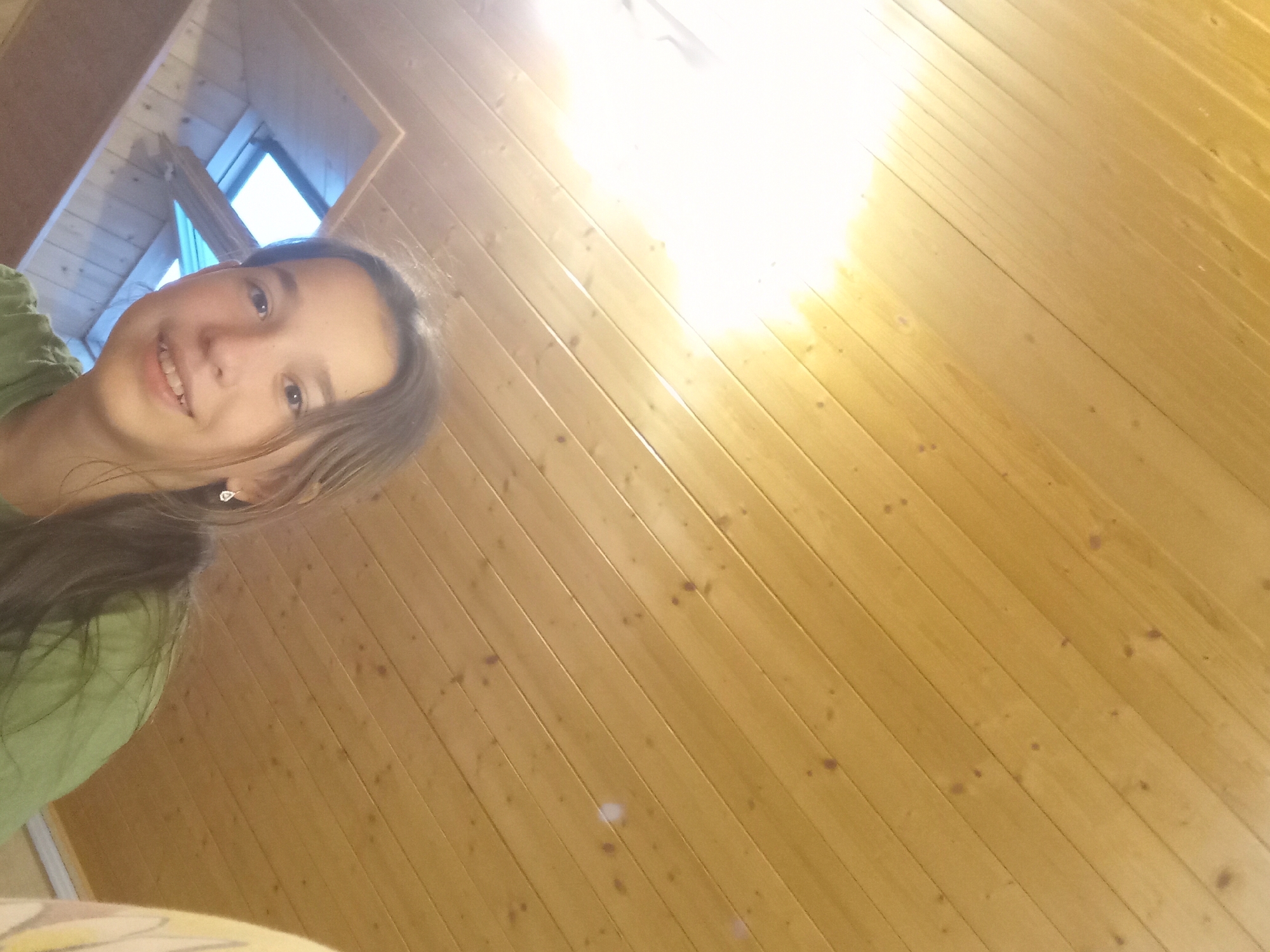 A young girl with shoulder-length brown hair and brown eyes smiles at the camera. She is wearing a green t-shirt and a small, dangling earring in her right ear. The background is a wooden-paneled ceiling with a skylight visible in the upper left portion of the image. The lighting suggests the image was taken indoors during the day.