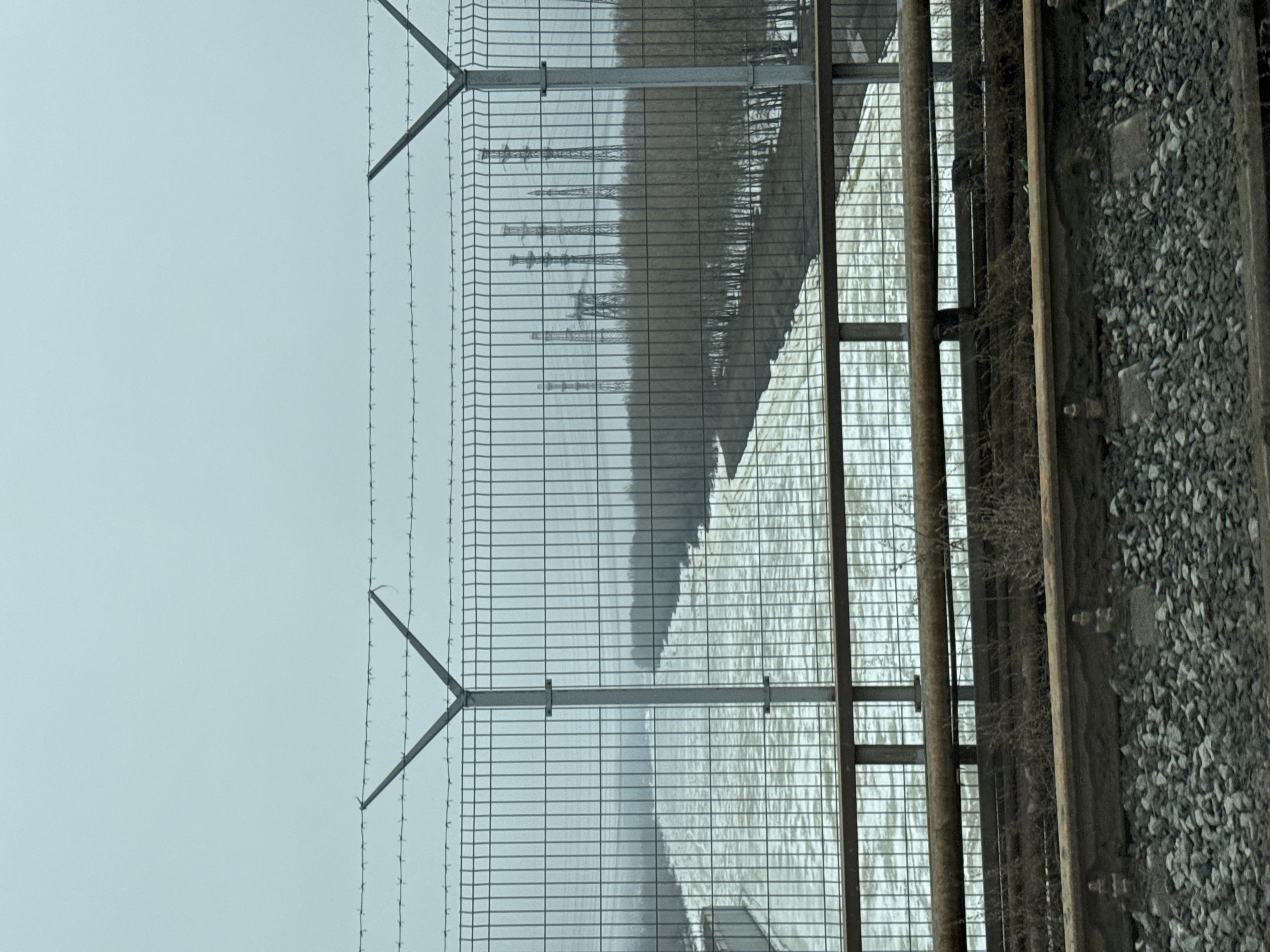 A metal fence with barbed wire stretches across the left side of the frame. Behind the fence, a blurry industrial scene with tall structures is visible. To the right of the fence, a metal structure stands adjacent to train tracks laid on a bed of grey gravel. The sky above is a pale, uniform grey. This is located within the city of Chaikovsky, Russia.
