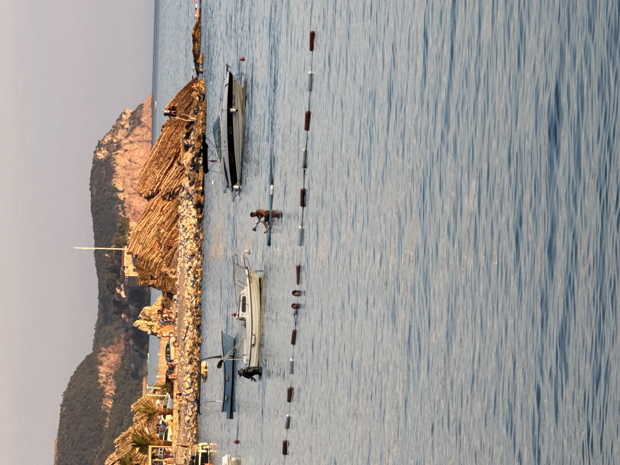 The image, taken in Rafailovići, Montenegro, depicts a calm body of water, likely the Adriatic Sea, with a textured surface reflecting the sunlight. On the left side of the frame, a rocky coastline is visible, adorned with thatched-roof structures that appear to be part of a beachfront establishment. A steep, rocky hill rises behind the coastline, partially covered in vegetation. Several boats are moored in the water. One is a small, light-colored motorboat with an outboard engine, positioned near the shore. Another, a dark-hulled boat, is further out, its mast visible. A person in a brown garment is in the water near a rope with buoys, seemingly engaged in an activity related to the boats or water. The scene suggests a peaceful day, likely late afternoon given the warm, golden light illuminating the rocks and water. The overall atmosphere is serene and typical of a coastal resort town.