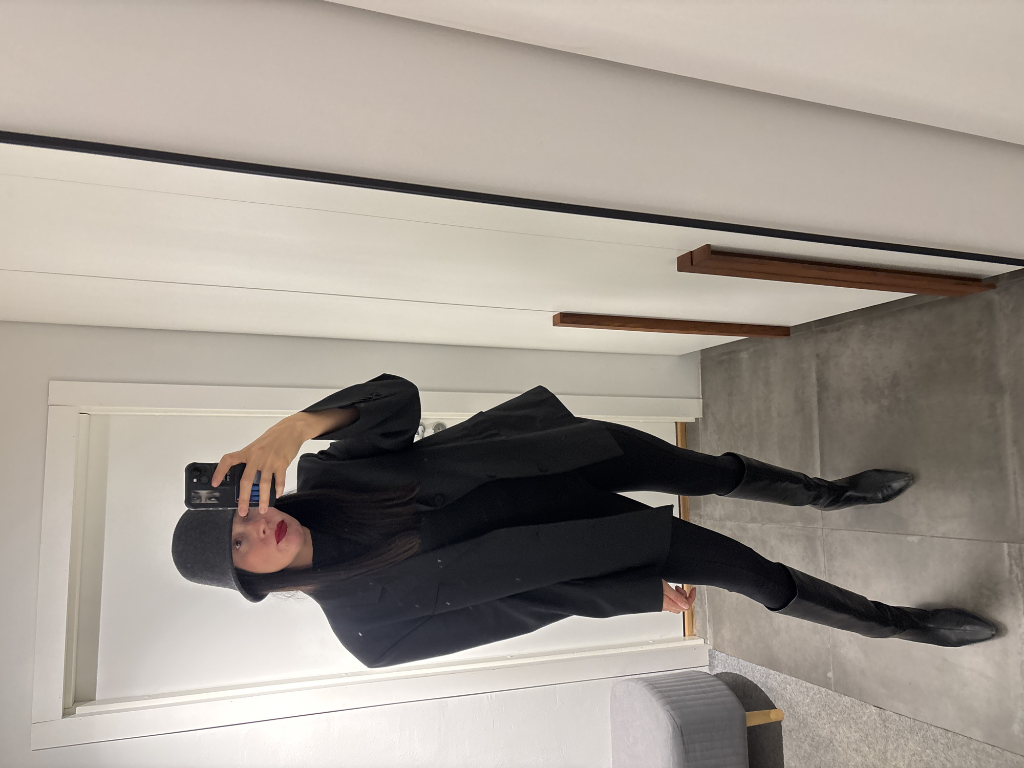 A woman is taking a selfie in what appears to be a dressing room or a walk-in closet, reflected in a full-length mirror. She is wearing a dark grey bucket hat, a black turtleneck sweater, black leggings or pants, a dark oversized blazer, and black knee-high boots. She is holding her phone in her right hand to take the picture. The room features white walls and a grey floor. There are wooden accents on the ceiling. A grey ottoman or bench is visible on the bottom left corner of the mirror's reflection. There's no clear indication in the image that the location is Chaikovsky, Russia.