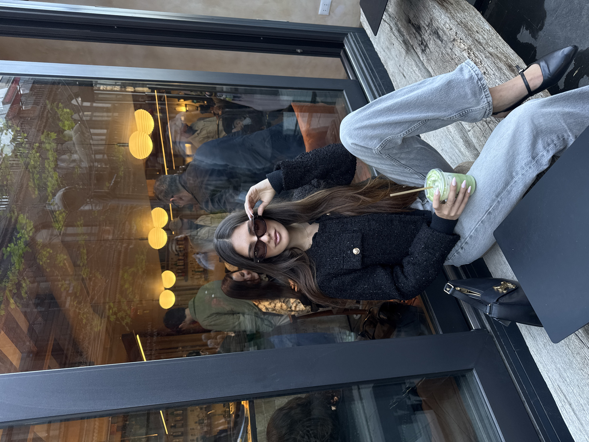 The image depicts a young woman sitting with her legs crossed at an outdoor cafe, likely in Yerevan, Armenia. She is wearing sunglasses, a dark, textured jacket with a gold button, and light-colored jeans. She holds a green beverage in a clear plastic cup with a straw. She sits on a wooden bench next to a black handbag and a black laptop. The background reveals a cafe interior with reflective glass, showing interior lighting fixtures and other patrons. The cafe is adjacent to a windowed storefront. The light suggests it is daytime.