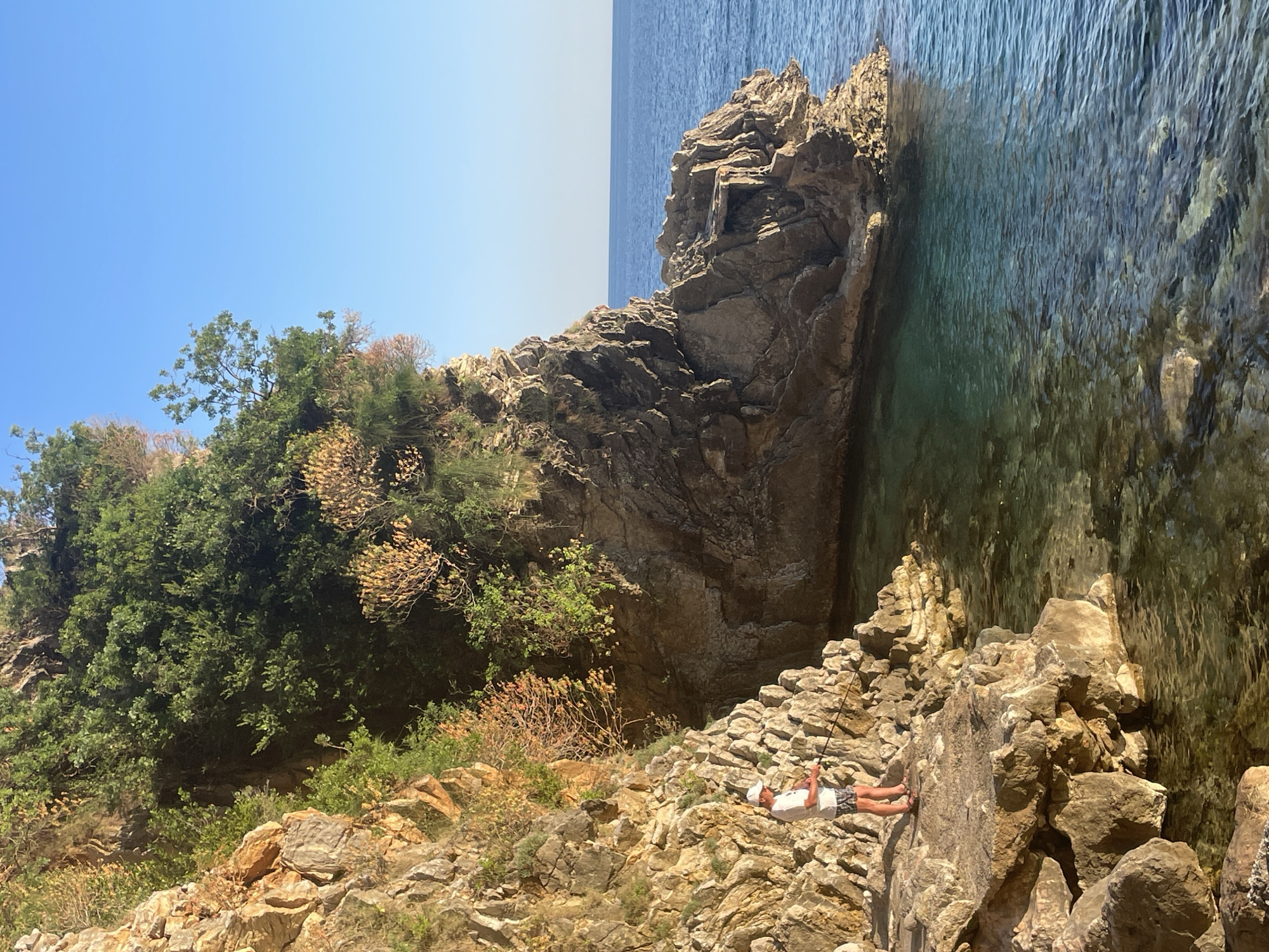 The image depicts a coastal scene in Boreti, Montenegro. The foreground features rocky terrain with a mix of sparse vegetation, including trees and shrubs. A person is lying on the rocks, appearing to be fishing with a rod. The middle ground showcases a large, rugged rock formation that juts out into the turquoise-colored water. The background consists of a clear blue sky and the horizon line where the sea meets the sky. The overall atmosphere is sunny and serene.