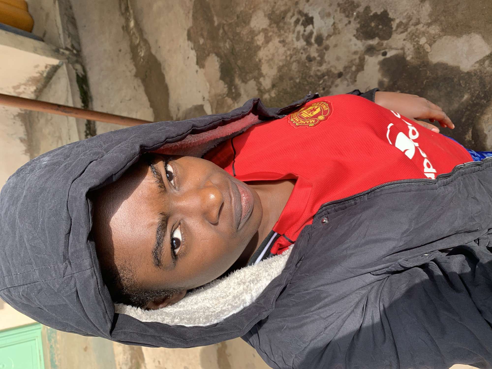 The image shows a close-up, high-angle, outdoor portrait of a young Black person wearing a black hooded jacket with a grey fuzzy lining and a red Manchester United football jersey. The individual's face is turned towards the camera, with their gaze directed slightly off-center. The background suggests an urban environment, with a dirty concrete ground, a textured wall, and a glimpse of a light green door or gate on the left. The lighting indicates it is daytime, with harsh shadows casting across the scene. The location is identified as Rafin Par, Nigeria.