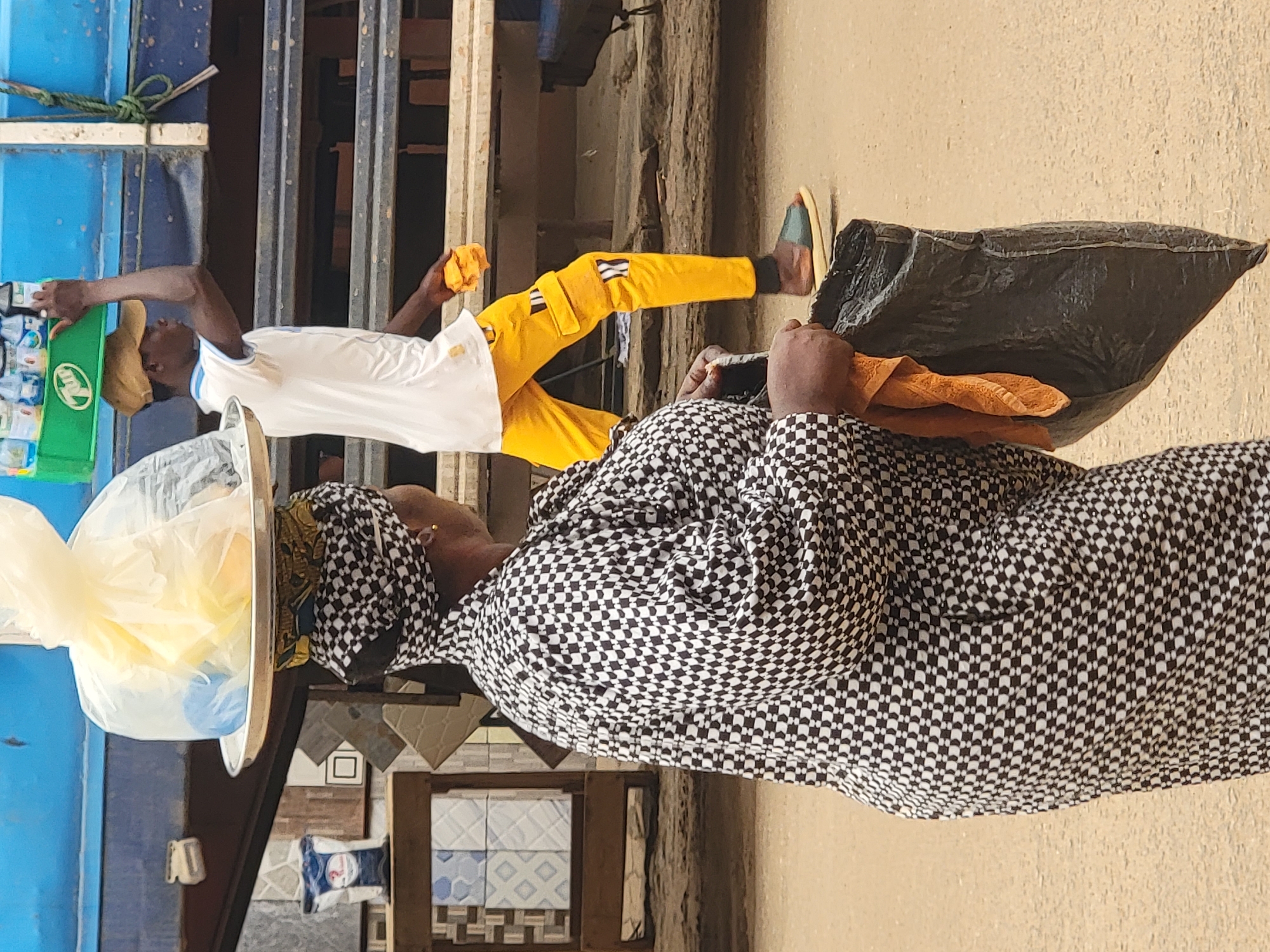 In Maiburiji, Nigeria, a woman in a black-and-white checkered outfit and headwrap stands outdoors. She holds a large black plastic bag containing an orange cloth. A young man wearing a white shirt, yellow pants with straps, and a tan cap appears to be buying something from her, reaching for a green package of water bottles. The backdrop includes a blue painted structure and a wooden doorway.