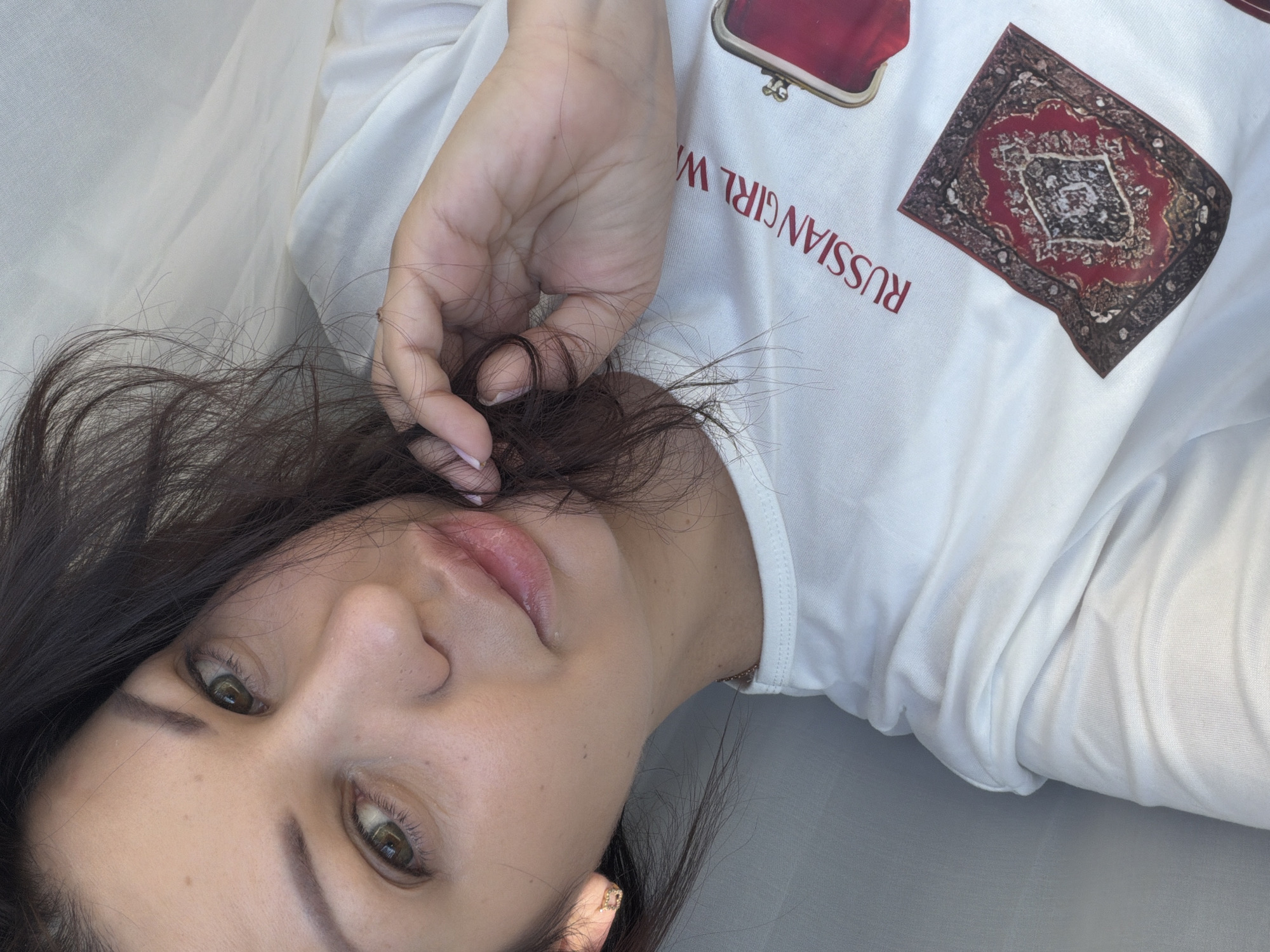 A young woman with dark hair and green eyes is lying down, possibly on a bed, with a white sheet draped around her. She is wearing a white t-shirt with a red graphic print that includes the words "RUSSIAN GIRL" and an image of a red coin purse, as well as a small Persian rug design. Her hand is near her face, and she is looking towards the camera. The lighting suggests it is daytime. The image is a close-up, focusing on the woman's face and upper torso. There is no discernible location within the city of Sochi, Russia, visible in the image.