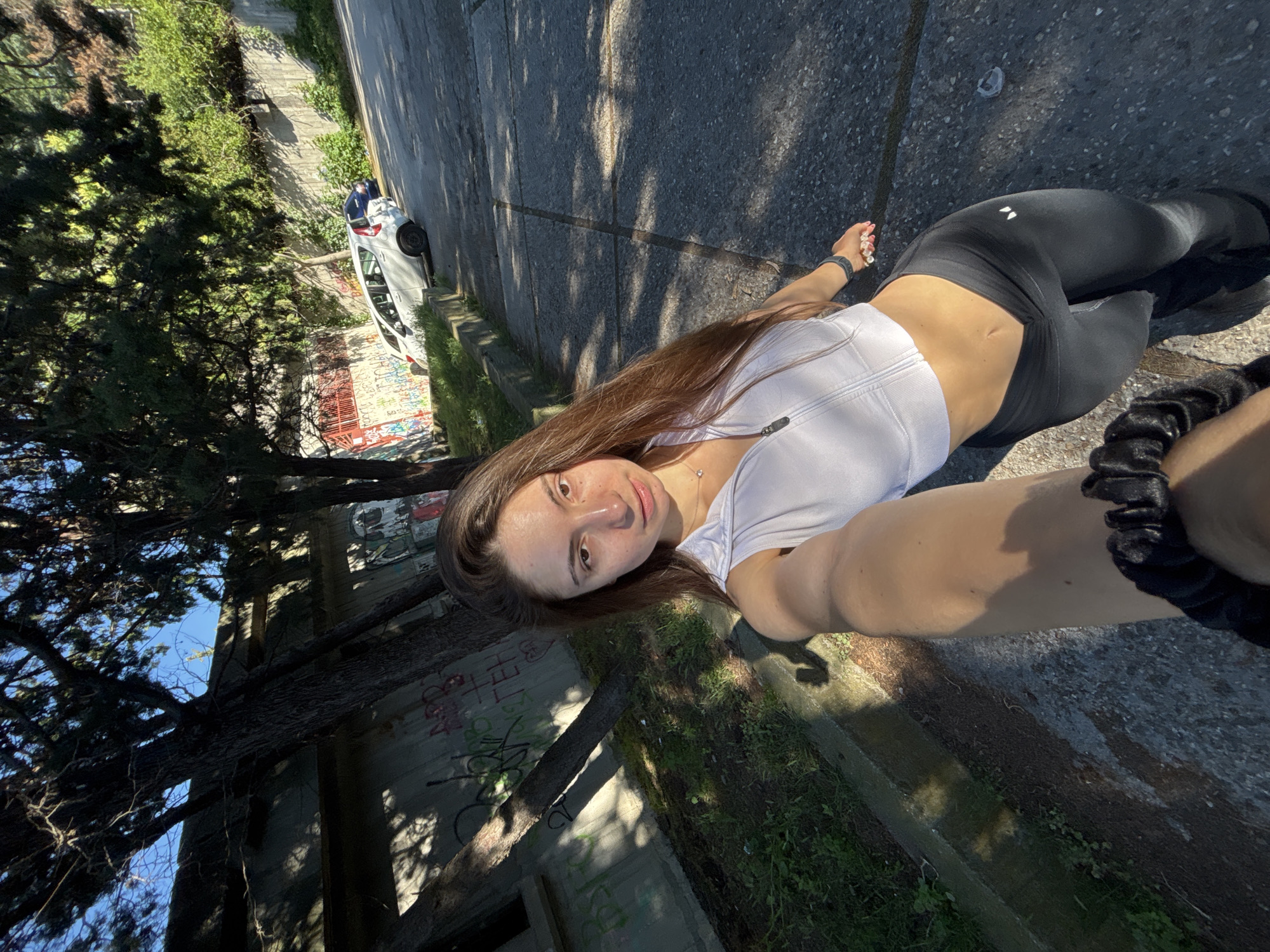 A woman is taking a selfie in an outdoor urban environment. She is wearing a white crop top and black leggings. The background shows a concrete wall with graffiti, trees, and parked cars. The lighting suggests it is daytime, with strong sunlight creating shadows. The location appears to be a street or pathway with a slight incline. There are no other people visible in the frame with the woman. The woman's pose and attire suggest she might be engaged in or preparing for physical activity.