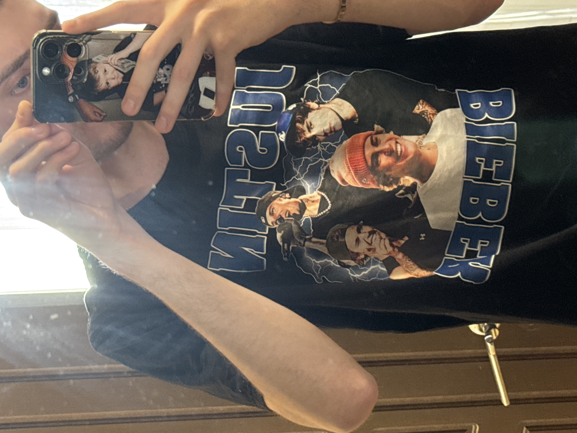 A person is taking a selfie in a mirror, wearing a black t-shirt with an image of Justin Bieber and the word "BIEBER" in blue. The phone in the person's hand has a case with an image of a child. The mirror reflection shows a door with a key inserted in the lock. The image does not contain any information that indicates this is happening in Tver, Russia.