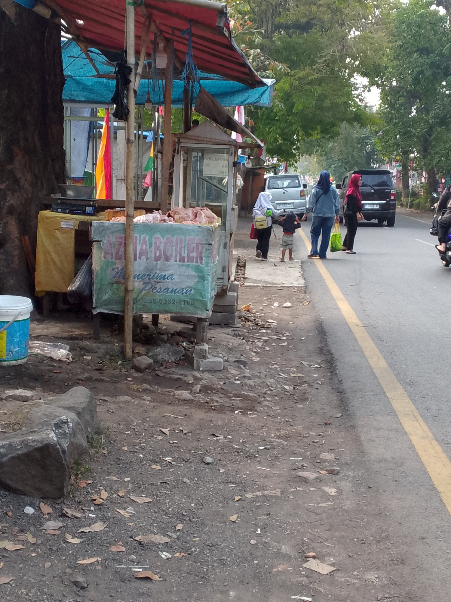 The image captures a street scene in Haurwangi, Indonesia, likely on a roadside. In the foreground, there's a stall with a red and blue awning, displaying raw meat on a counter. The stall has green and white signage that reads "AZIZIA BOILER" and "Menerima Pesanan" with a phone number "0895-0362-2190". To the left of the stall, a blue bucket and other debris are visible on the ground, which is mostly dirt and scattered leaves.
Further down the street, several people are present. Two women, one in a blue denim jacket and headscarf and another in a red headscarf, walk on the road. A child in a grey shirt and shorts walks between them. A white car is parked on the left side of the road, and a black SUV is driving on the right lane. The road itself has a prominent yellow line marking the lanes. Trees and foliage line the background, suggesting a moderately vegetated urban or semi-urban environment. The overall atmosphere appears to be a typical daytime scene in a local market area or along a busy street.