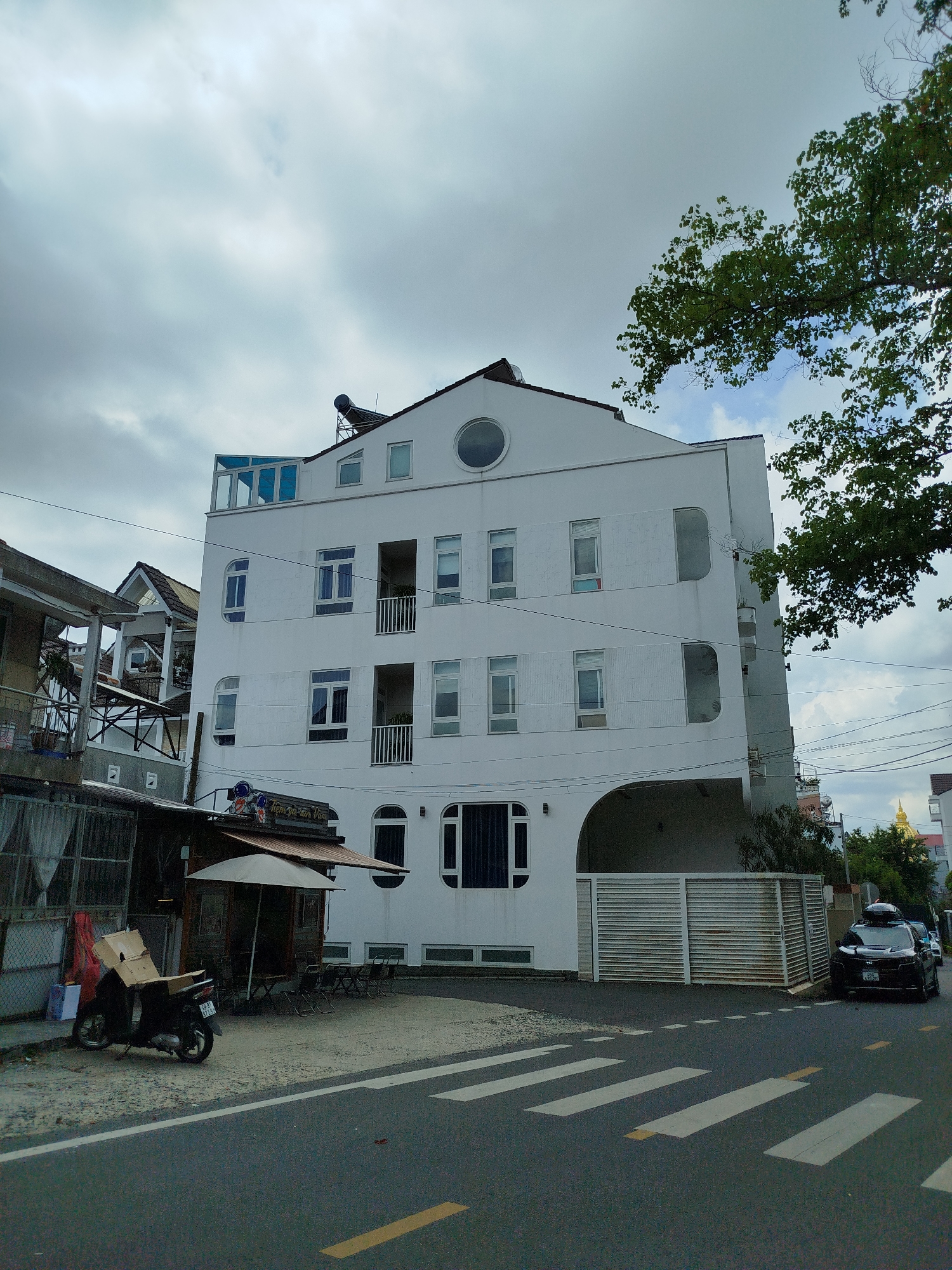 In Phường Xuân Hương, Đà Lạt, Vietnam, a multi-story white building with a distinctive curved entrance is prominently featured. The building has numerous rectangular windows of varying sizes, a circular window on the upper level, and a glass-enclosed structure on the rooftop. A solar water heater is visible on the roof. Adjacent to the white building, another structure with a pitched roof and balconies is partially visible. In front of the white building, a small covered seating area with umbrellas and chairs is set up, and a dark-colored scooter with a cardboard box strapped to its back is parked on the sidewalk. The street in the foreground has a crosswalk marked with white and yellow lines. A black SUV with a roof rack is parked near a slatted metal gate. The sky is overcast with scattered clouds.