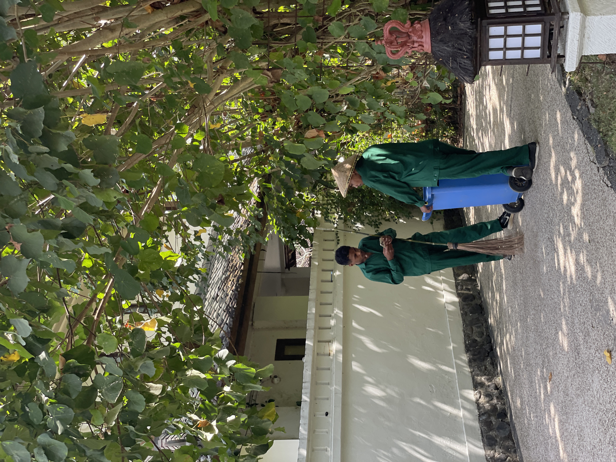 Two men in green uniforms are standing on a paved pathway lined with a white wall and a stone base. The man on the left is looking at his phone, holding a broom in his right hand. The man on the right, wearing a conical hat, is standing next to a blue trash can with wheels. The scene is outdoors, shaded by lush green trees with dappled sunlight falling on the ground. In the background, a building with a tiled roof is visible behind the white wall. The overall environment suggests a well-maintained outdoor area, possibly a resort or a public space in Kuta Selatan, Indonesia.