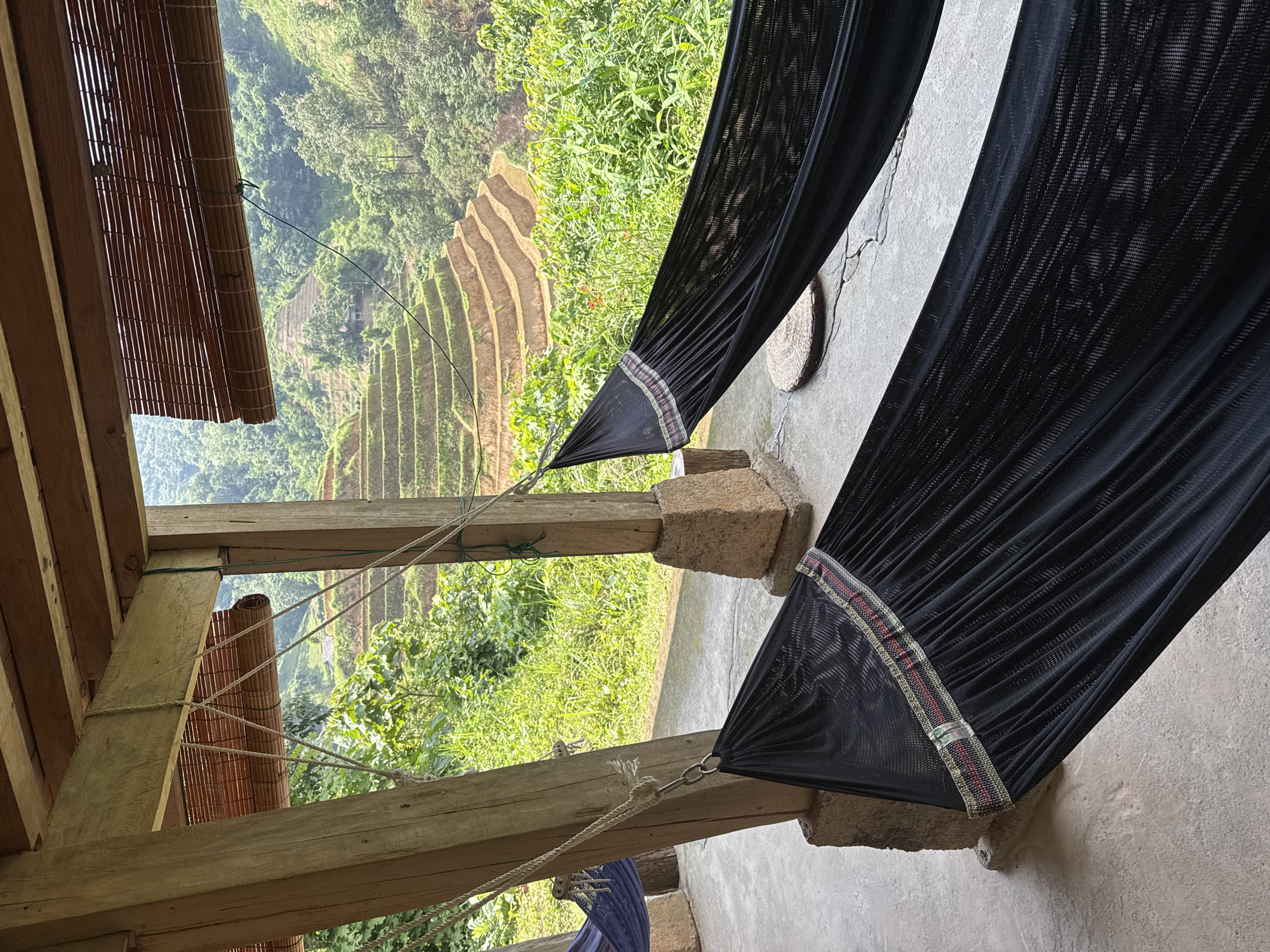 A hammock is suspended between the wooden pillars of a structure, potentially a porch or open-air building, overlooking the terraced rice paddies in the Hoàng Su Phì Commune, Vietnam. The hammock is made of black mesh fabric with a decorative band at the ends. The support pillars are of weathered wood and stone. A bamboo roll-up blind is partially visible. The terraced fields are filled with green vegetation. The landscape appears mountainous and lush with trees and other plants. The sky is overcast, suggesting a cloudy or humid day.