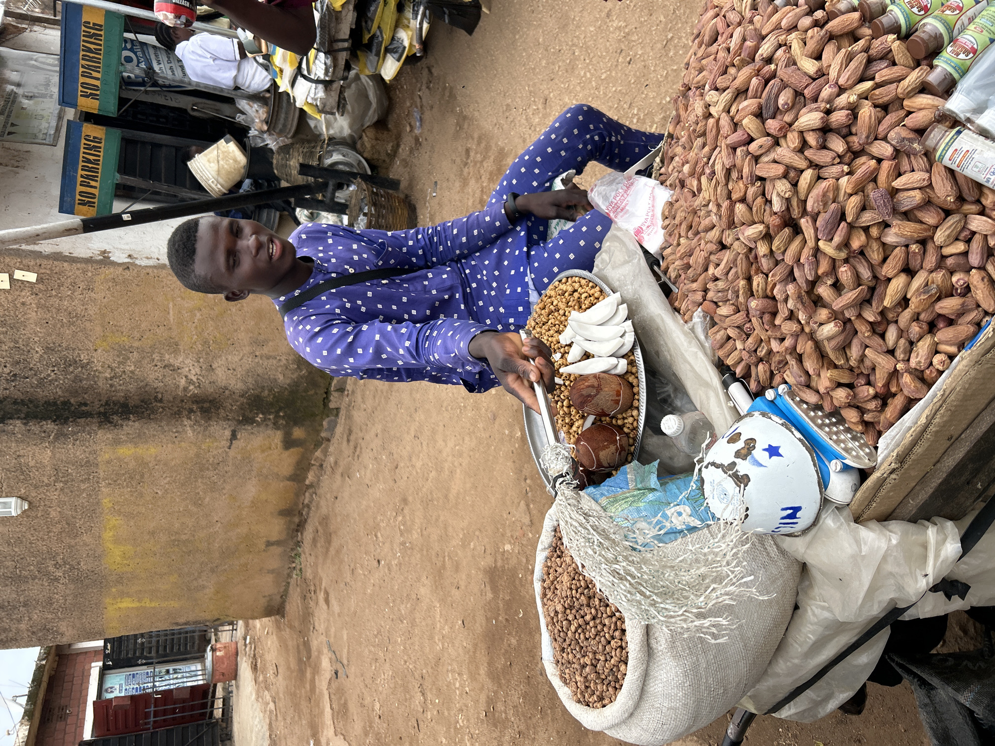 A young man sits in a blue and white patterned outfit, holding a metal tray with two round, brown objects, sliced white fruit, and small nuts. He is positioned next to a large pile of dried dates and other goods. The scene appears to be an outdoor market, with a dirt ground and a rough, textured wall in the background. Several signs are visible, including two that read "NO PARKING" in English. There are also several bottles of what appear to be spices or condiments, one labeled "VIVA POLY BA" and another with "SASANG". The man's expression is neutral. The lighting suggests it is daytime, but the weather is unclear. The overall impression is a bustling market stall in an unknown location.