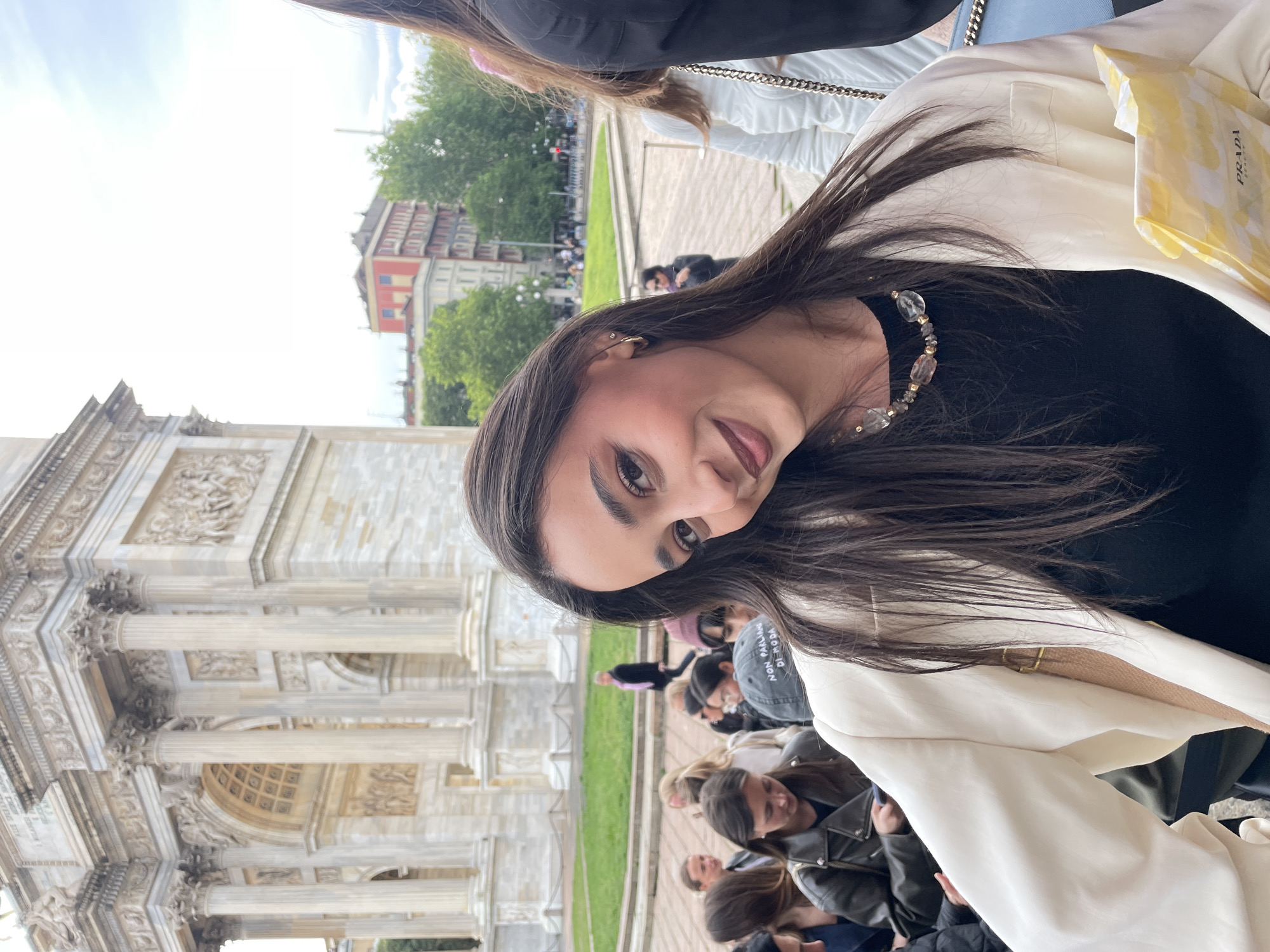 A young woman is posing for a photo in front of the Arco della Pace in Milan, Italy. The woman has long brown hair, is wearing a black shirt with a beaded necklace, and a white blazer. She is holding a Prada shopping bag. Behind her, a crowd of people is walking in the piazza, and in the background, there are trees and buildings. The weather appears to be overcast. The architecture of the Arco della Pace is visible, with its detailed carvings and columns.