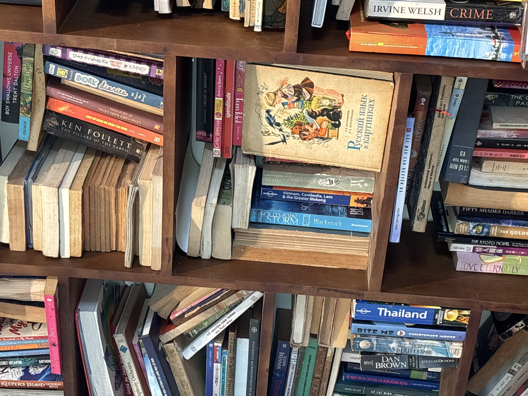 The image shows a bookshelf filled with various books in different languages, including English, Vietnamese, and Russian. The books cover a range of genres, from fiction and travel guides to textbooks. Titles like "Boy Swallows Universe," "Dork Diaries," "Ken Follett The Pillars of the Earth," "Crime" by Irvine Welsh, "Thailand," and "Dan Brown Angels & Demons" are visible. There's also a book titled "Русский язык в картинках" (Russian Language in Pictures). The books are densely packed and arranged somewhat haphazardly on the shelves. There's no clear indication of the location being Đà Nẵng, Vietnam, beyond the presence of Vietnamese books. The shelf appears to be part of a home or private library, given the diverse and personal collection of books.