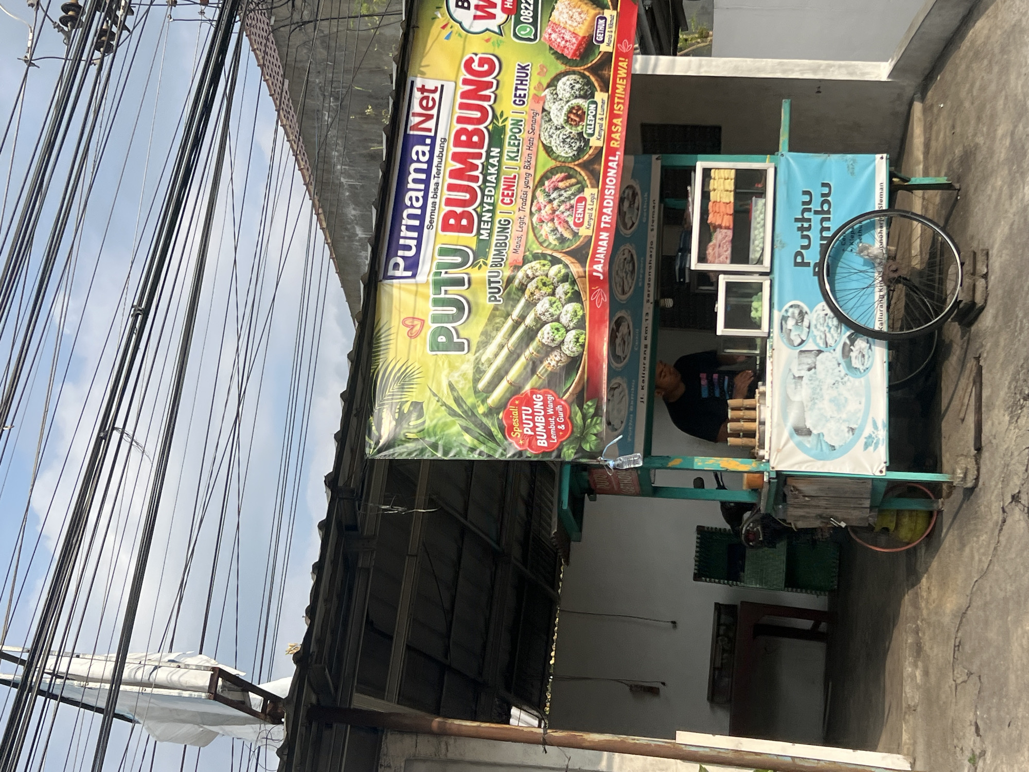 The image depicts a street-side food stall in Ngaglik, Indonesia, selling traditional Indonesian sweets. The stall is a mobile cart with a teal-colored frame and a large, vibrant banner advertising "Purnama.Net PUTU BUMBUNG." The banner showcases various treats: Putu Bambung, Cenil, Klepon, and Gethuk, with descriptions of their flavors and textures. Next to the banner, a portion of the cart displays colorful boxes filled with these sweets. In the background, a person is partially visible behind the counter, appearing to be attending to customers or preparing food. A motorcycle is parked behind the stall, and various household items and a gas cylinder are visible in the surrounding area, suggesting a residential or mixed-use neighborhood. Overhead, a complex network of power lines traverses the sky, characteristic of many urban and semi-urban settings in Indonesia. The ground is paved with concrete, showing signs of wear and tear. The overall scene is one of everyday commerce on a street in Indonesia.