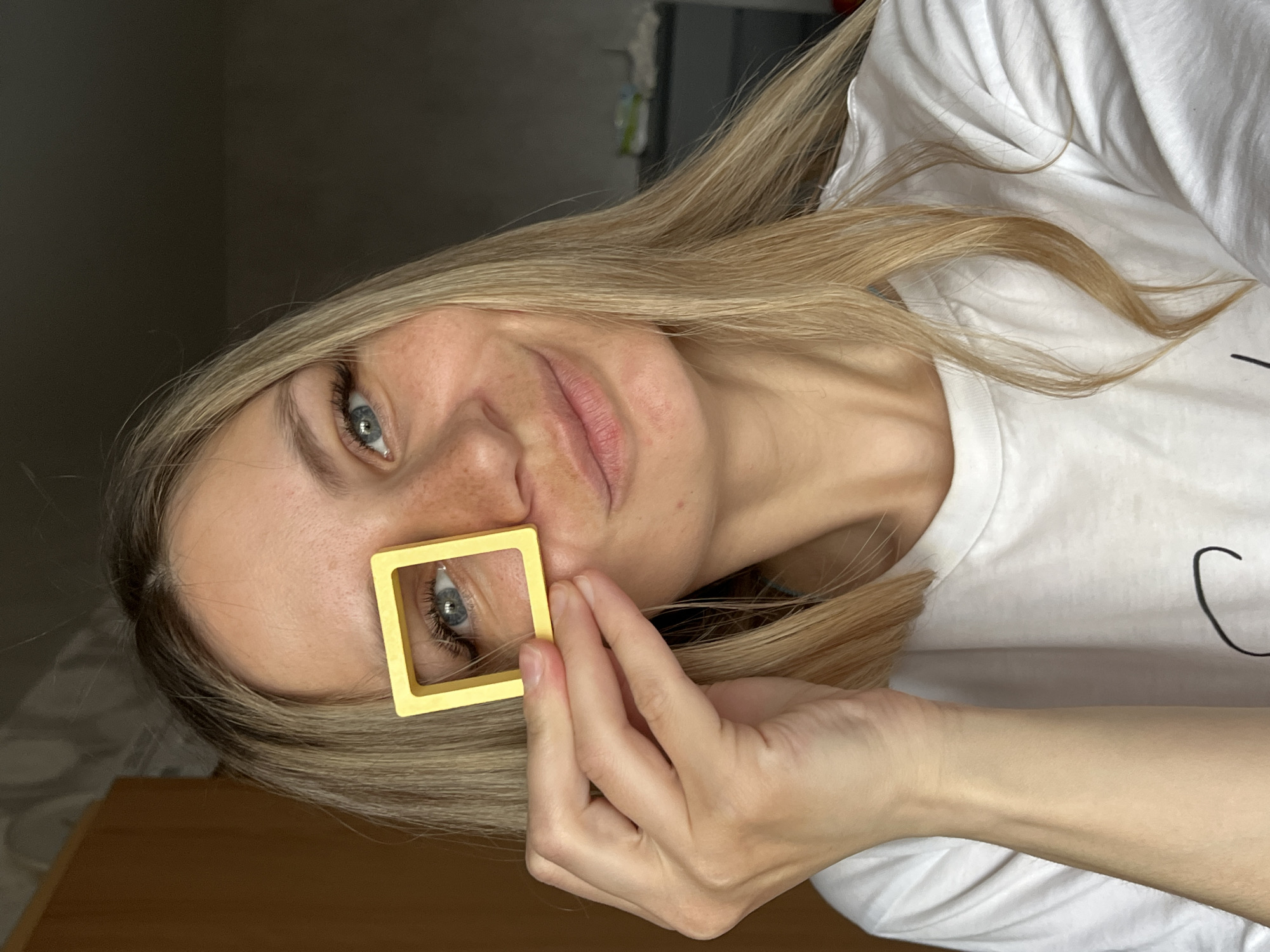 A woman with blonde hair and blue eyes is holding a yellow square frame up to her left eye. She is wearing a white t-shirt. The background is a dimly lit room with a wooden table visible in the lower left corner. It is not possible to confirm the location in the city of Chaikovsky, Russia from the image alone.