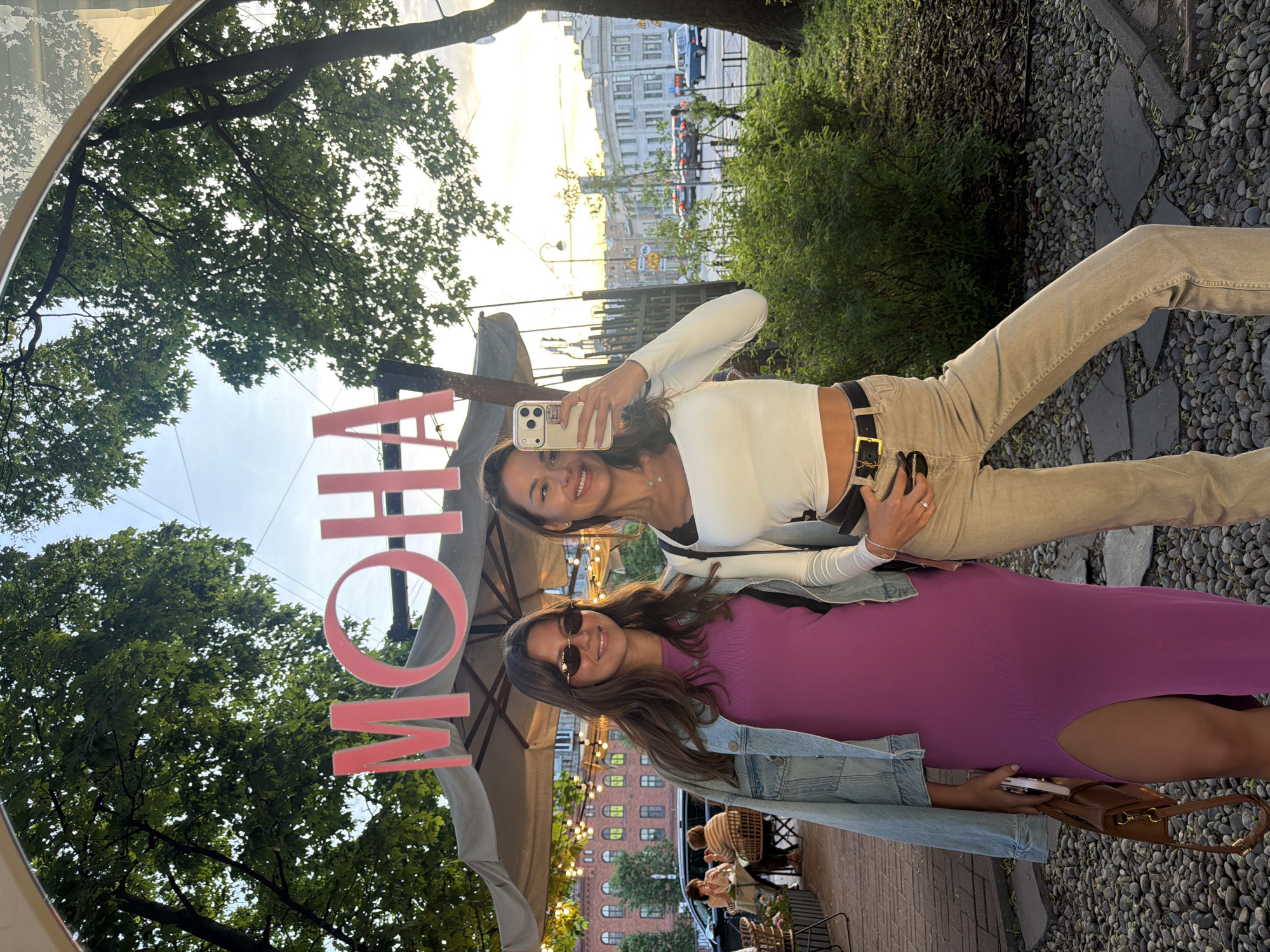 Two young women pose for a mirror selfie in front of a cafe in Saint Petersburg, Russia. The cafe is decorated with pink lettering that spells "MOHA" in the reflection. The woman on the left wears a purple dress and a denim jacket, while the woman on the right sports a white crop top and beige pants. They stand on a cobblestone path, with lush green trees and a cityscape visible in the background. The city skyline features classic European architecture, and a street with cars can be seen in the distance. The scene appears to be set during the golden hour, with warm sunlight illuminating the surroundings.