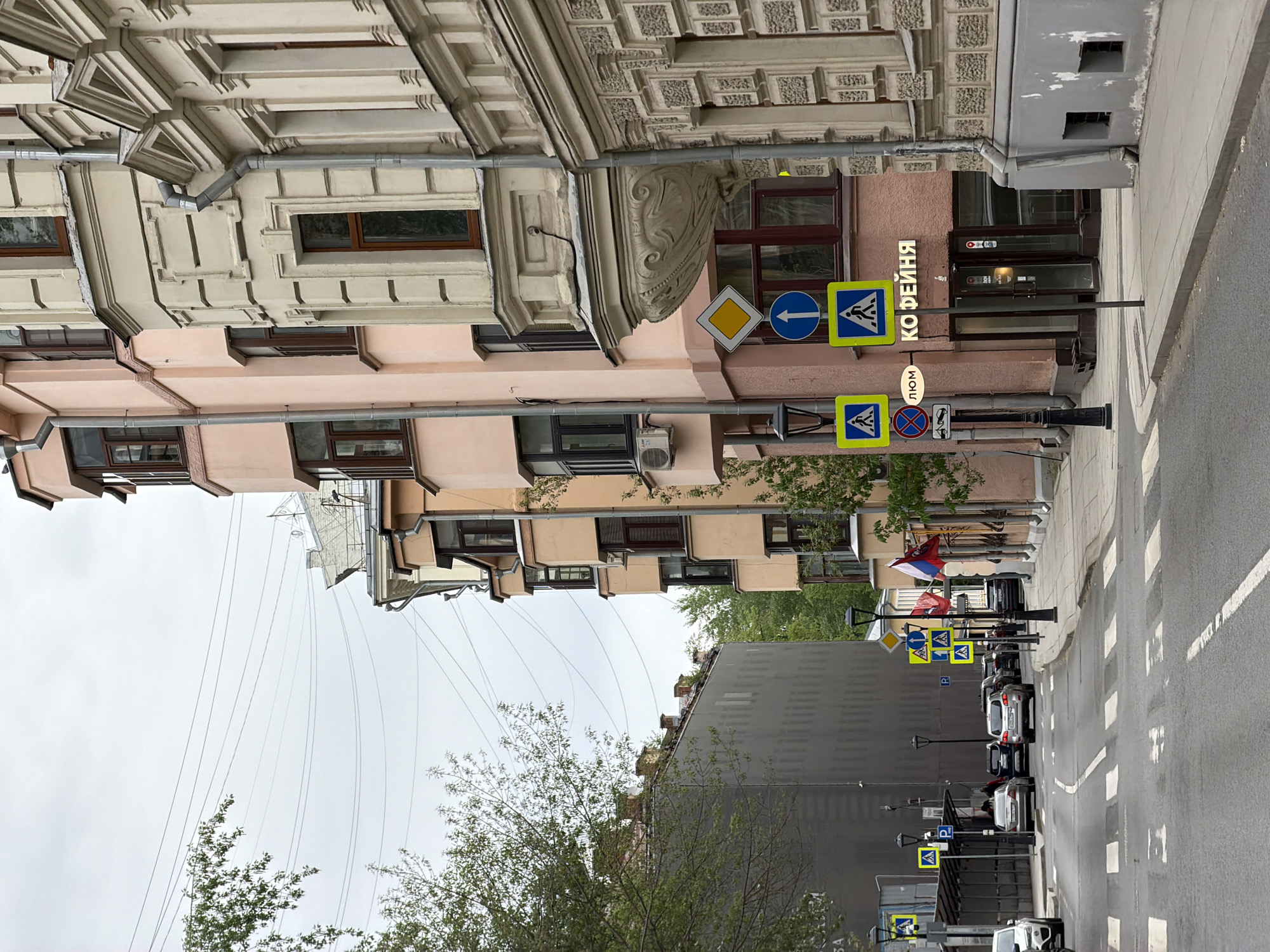 The image depicts a street in Moscow, Russia. The architecture is characterized by multi-story buildings with ornate facades, including intricate moldings and varied window styles. A pedestrian crosswalk is visible on the right, with zebra stripes marking the pavement. Several traffic signs are mounted on posts along the sidewalk, including signs for pedestrian crossings, directions, and parking regulations. Cars are parked along the side of the road. A street light and trees add to the urban landscape. The sky is overcast, suggesting a cloudy day. A building facade is adorned with Russian flags. A store sign that reads "КОФЕЙНЯ" in Russian, which translates to "Coffee Shop".