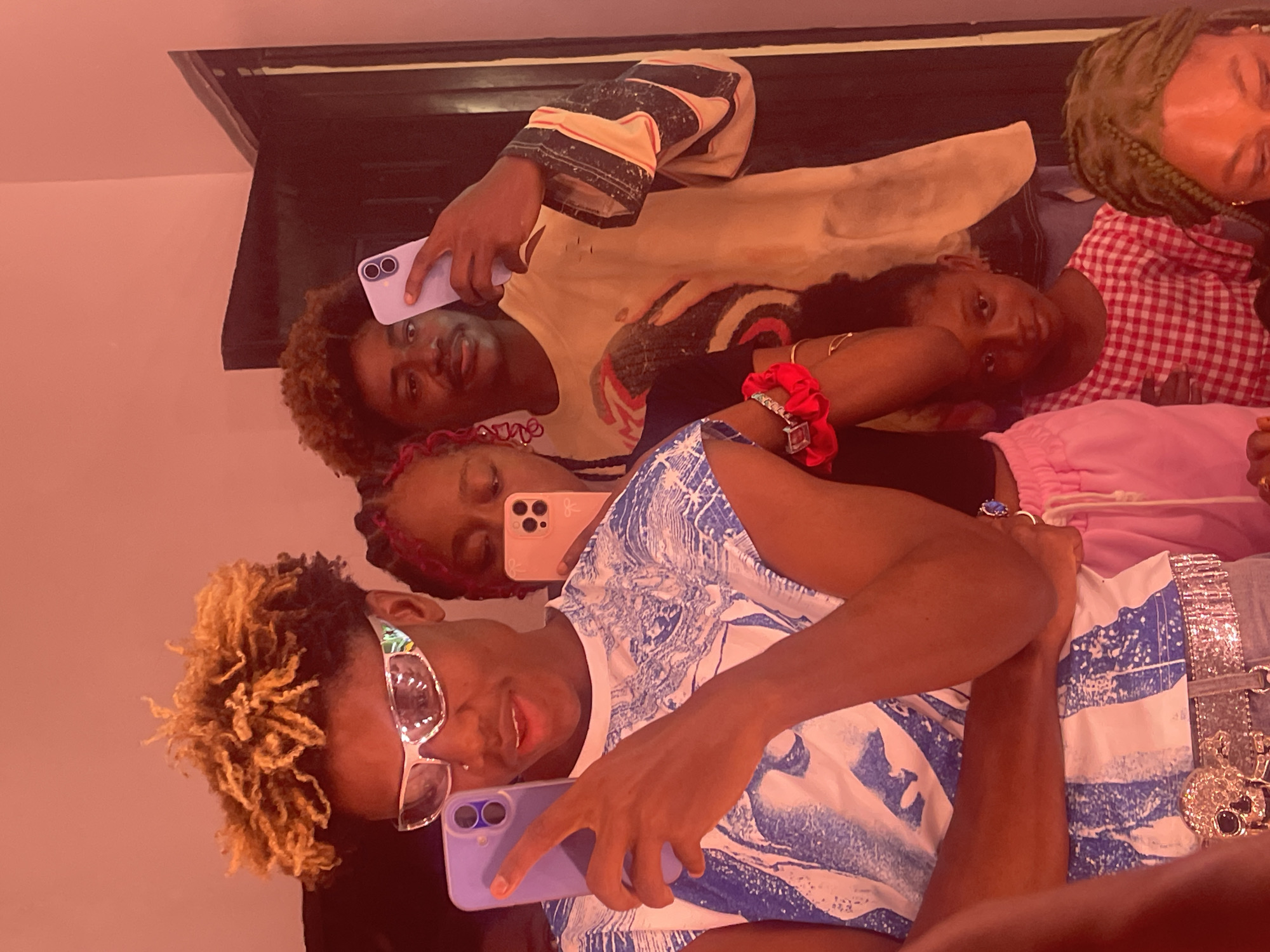 The image shows a group of five people indoors, possibly taking a selfie or group photo. The location is identified as Igboefon, Nigeria, based on the prompt. The person in the foreground wears a white and blue patterned shirt, silver sunglasses, and appears to be holding a purple smartphone. Another person is wearing a pink head wrap, black shirt, and pink pants, also holding a pink smartphone. A third person wears a beige shirt with black and white designs. Another person is wearing a red and white checkered shirt. The fifth person is partially visible with green hair. All individuals appear to be positioned closely together, suggesting a social gathering or casual setting. The background is indistinct, with a dark upper section and a lighter-colored wall visible on one side.