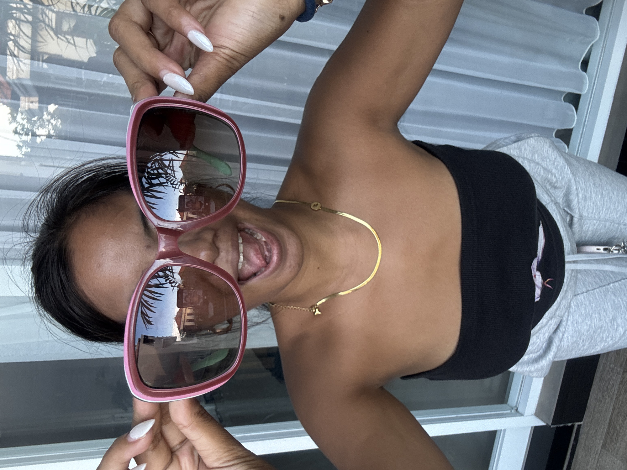 The image captures a close-up, rotated portrait of a woman engaged in a playful activity. She holds a pair of large, pink-framed sunglasses up to her face, partially obscuring her eyes. Her mouth is wide open, suggesting a laugh or a broad, joyful smile. Her hands display long, pointed nails painted in a light, possibly white or iridescent, color.
The woman is wearing a black strapless top or bandeau, a thin gold-colored choker necklace with a small pendant, and what appear to be grey sweatpants. A blue wristband or bracelet is visible on her right arm.
The background shows what appears to be an interior or sheltered outdoor space with white window frames or blinds behind her. Notably, the lenses of the sunglasses reflect an outdoor scene, revealing palm tree fronds and parts of a building, indicative of a sunny, tropical setting consistent with Kuta, Indonesia. The time of day is clearly daytime. The overall impression is one of casual happiness and playfulness. There is no visible text in the image.