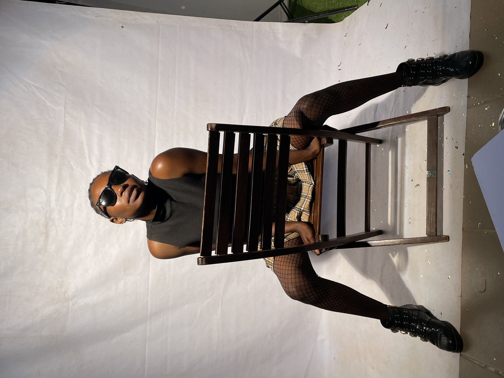 The image depicts a person sitting on a wooden chair in a studio setting. The person is wearing sunglasses, a dark sleeveless top, a plaid skirt, fishnet stockings, and black boots with metal accents. The studio has a white backdrop with some confetti scattered on the floor. The location in the city of Jos, Nigeria, is not evident in the image.