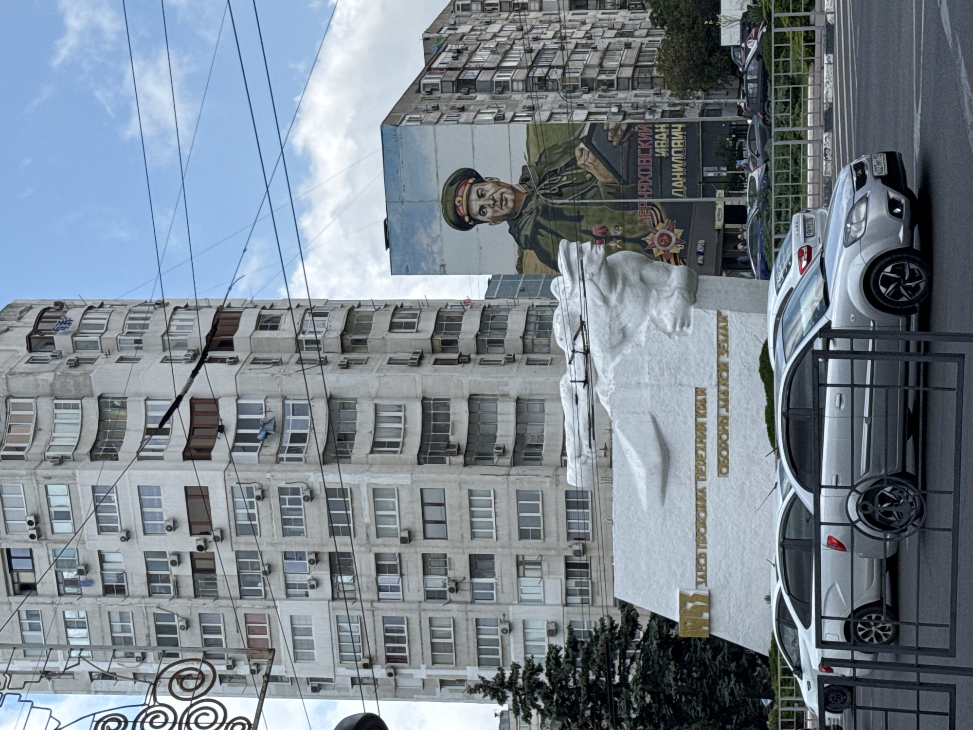 This media file captures a street scene in Novorossiysk, Russia. The primary subjects include a large mural on the side of a tall, weathered apartment building depicting a man in a military uniform. The mural features Cyrillic text above the man's head. In front of the apartment building and mural is a white, abstract sculpture or monument with more Cyrillic text visible on its base. Parked along the street are several white cars, one of which is prominently featured in the foreground. The scene appears to be outdoors during the daytime, with a cloudy sky. The overall impression is an urban environment with architectural elements characteristic of Soviet-era construction, juxtaposed with a modern monument and street-level activity. The presence of the mural and monument suggests a commemoration or memorial of some kind.