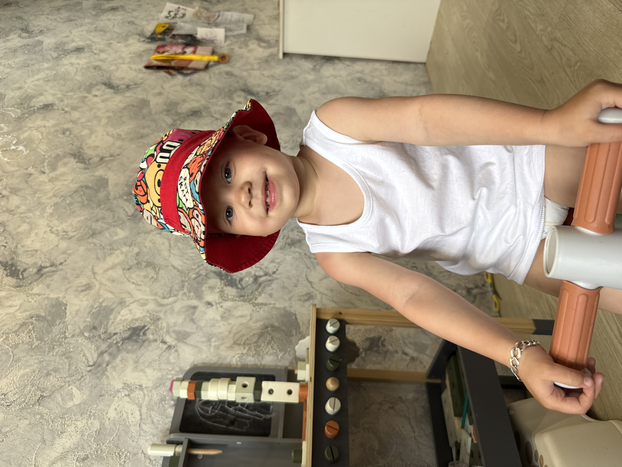 A young child wearing a white tank top and a colorful bucket hat is smiling at the camera. The child is standing next to a wooden play structure with various tools and knobs. The background shows a patterned floor and a cabinet. The location, Rusanovo, Russia, is not determinable from the image.