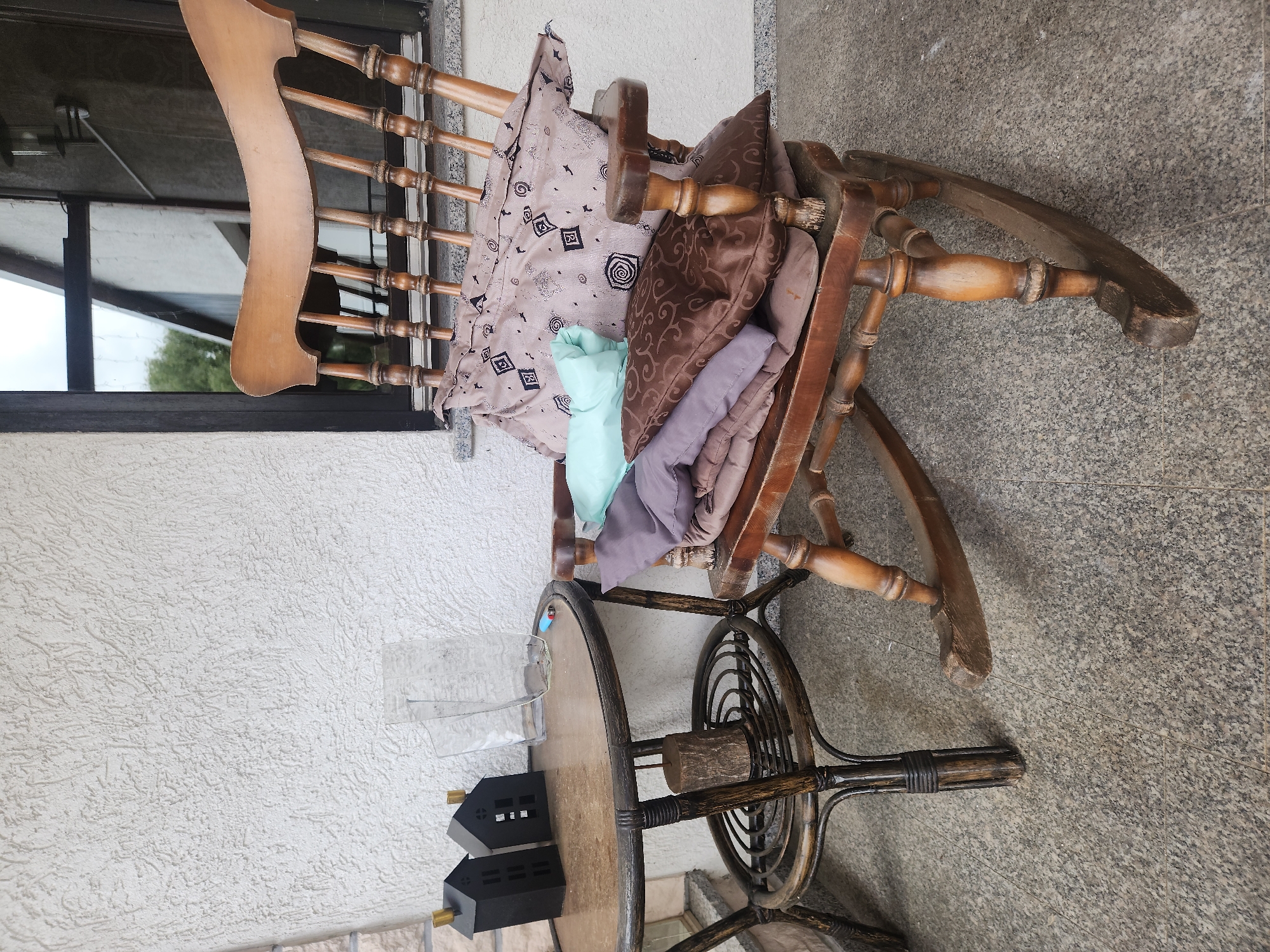 A vintage wooden rocking chair is placed on a granite tile floor, possibly on a porch or patio. The chair is adorned with several cushions, including one with a patterned beige cover, a light blue one, and two in muted purple and brown tones. To the left of the chair, a small, round wicker table with a glass top holds two miniature black house decorations and a clear glass vase. The background features a textured white wall and a window. The scene suggests a relaxed outdoor setting.