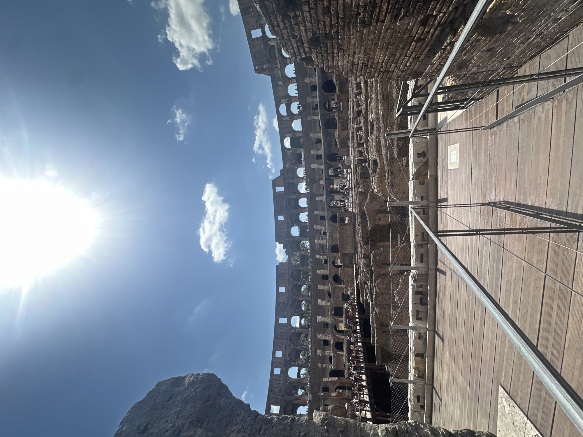 The image shows a view of the Colosseum in Rome, Italy. The sun is bright in the sky, with some clouds present. The Colosseum's ancient stone structure is visible, with people walking on a wooden walkway in the foreground. The walkway has metal railings for safety. The background shows the interior of the Colosseum, with its tiered seating and exposed underground structures. There are many tourists visible on the different levels of the amphitheater. The overall scene depicts a sunny day at a historical landmark.