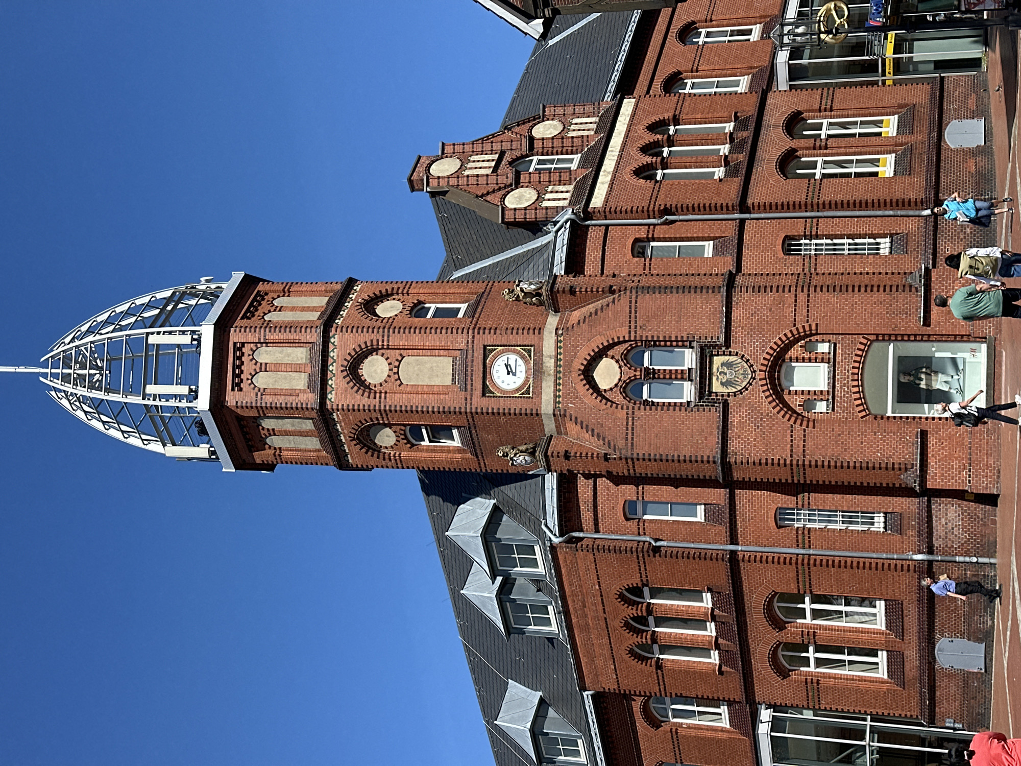 The image shows the Stade Town Hall in Stade, Germany, a red brick building with a distinctive clock tower topped by a modern glass dome. The sky is clear and blue. Several pedestrians are visible walking on the sidewalk next to the building. Some storefronts are visible at the building's base.