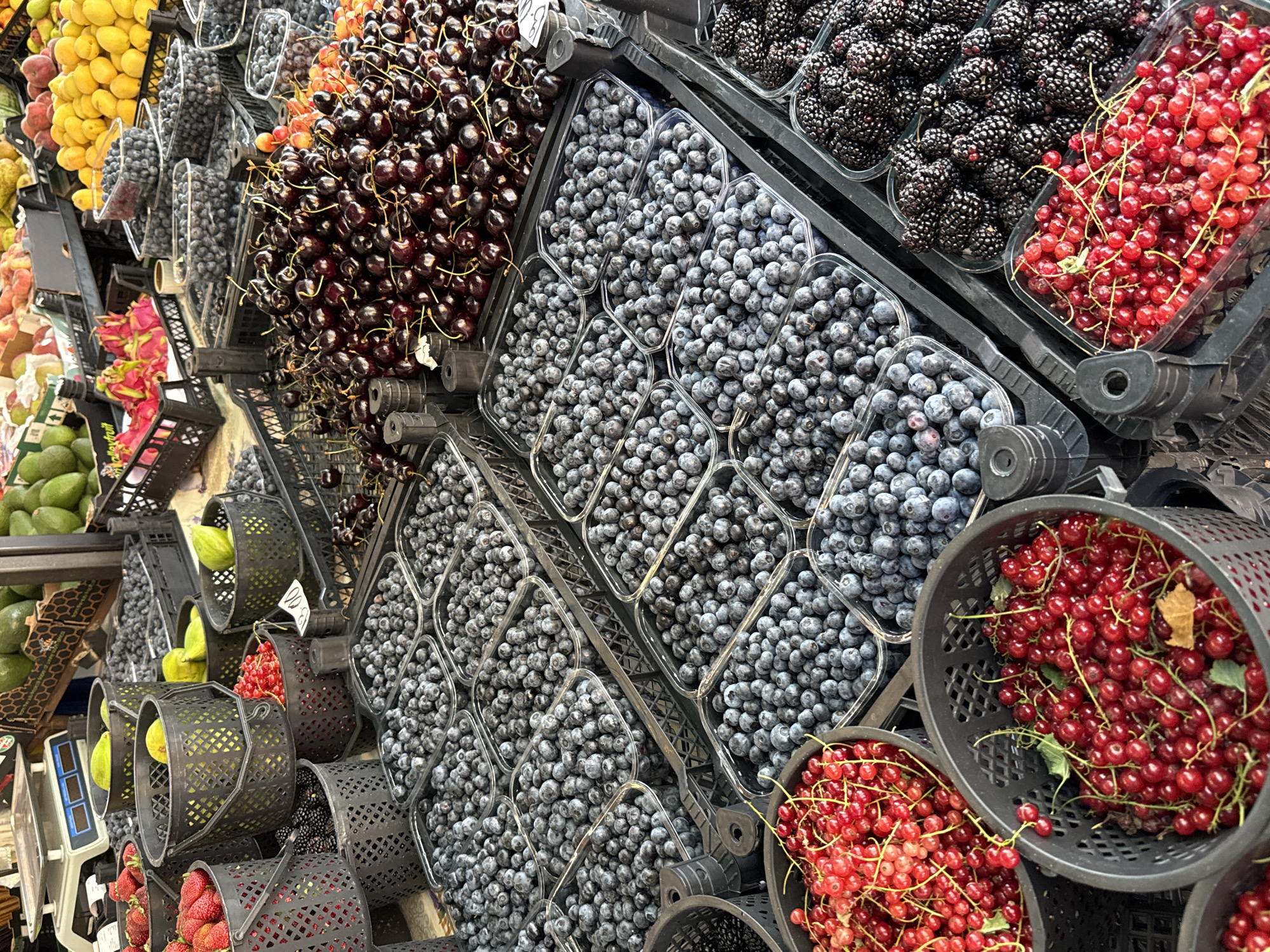The image displays a close-up view of a fruit stall at an outdoor market. Numerous baskets and containers are filled with a variety of fresh fruits, including strawberries, blueberries, cherries, red currants, and blackberries. The fruits are meticulously arranged, showcasing their vibrant colors and textures. The stall appears to be well-stocked, suggesting a bountiful harvest. The overall impression is one of freshness and abundance, with the fruits inviting consumers to sample their natural sweetness. The setting is likely a bustling marketplace, alive with the energy of commerce and the aroma of fresh produce.