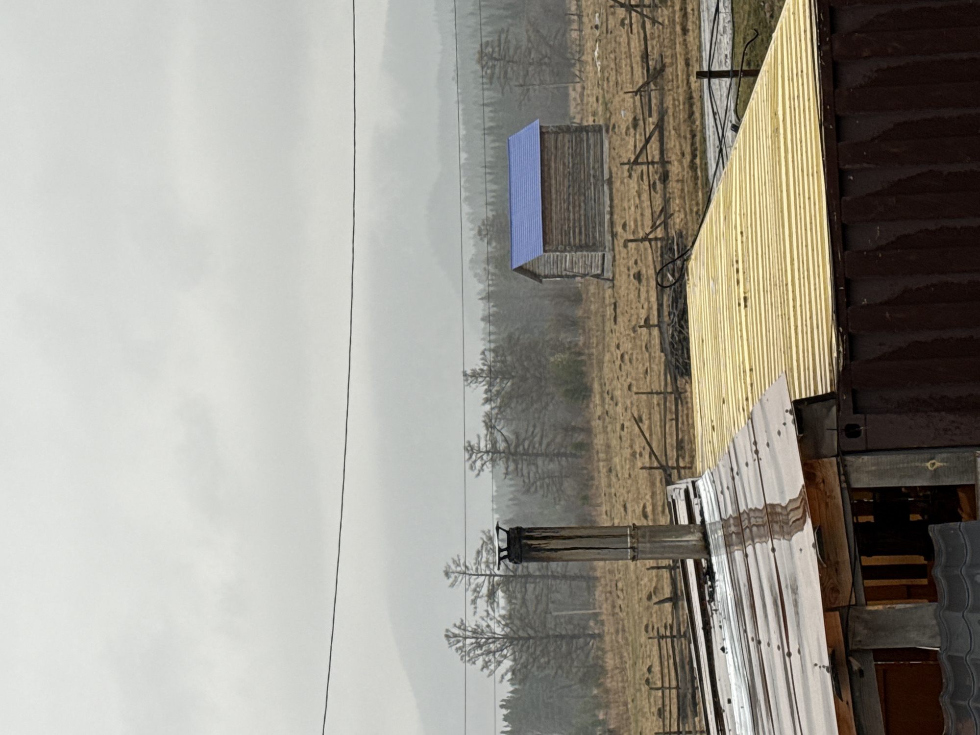 The scene depicts a rural landscape in Tyulyuk, Russia, under a cloudy sky suggesting precipitation. A small, weathered wooden structure with a blue roof stands in a field with sparse vegetation. A wooden fence partially encloses the area. In the foreground, parts of a corrugated metal building are visible, with a chimney extending upwards. Power lines stretch across the sky. The environment is foggy, obscuring distant mountains and forests.