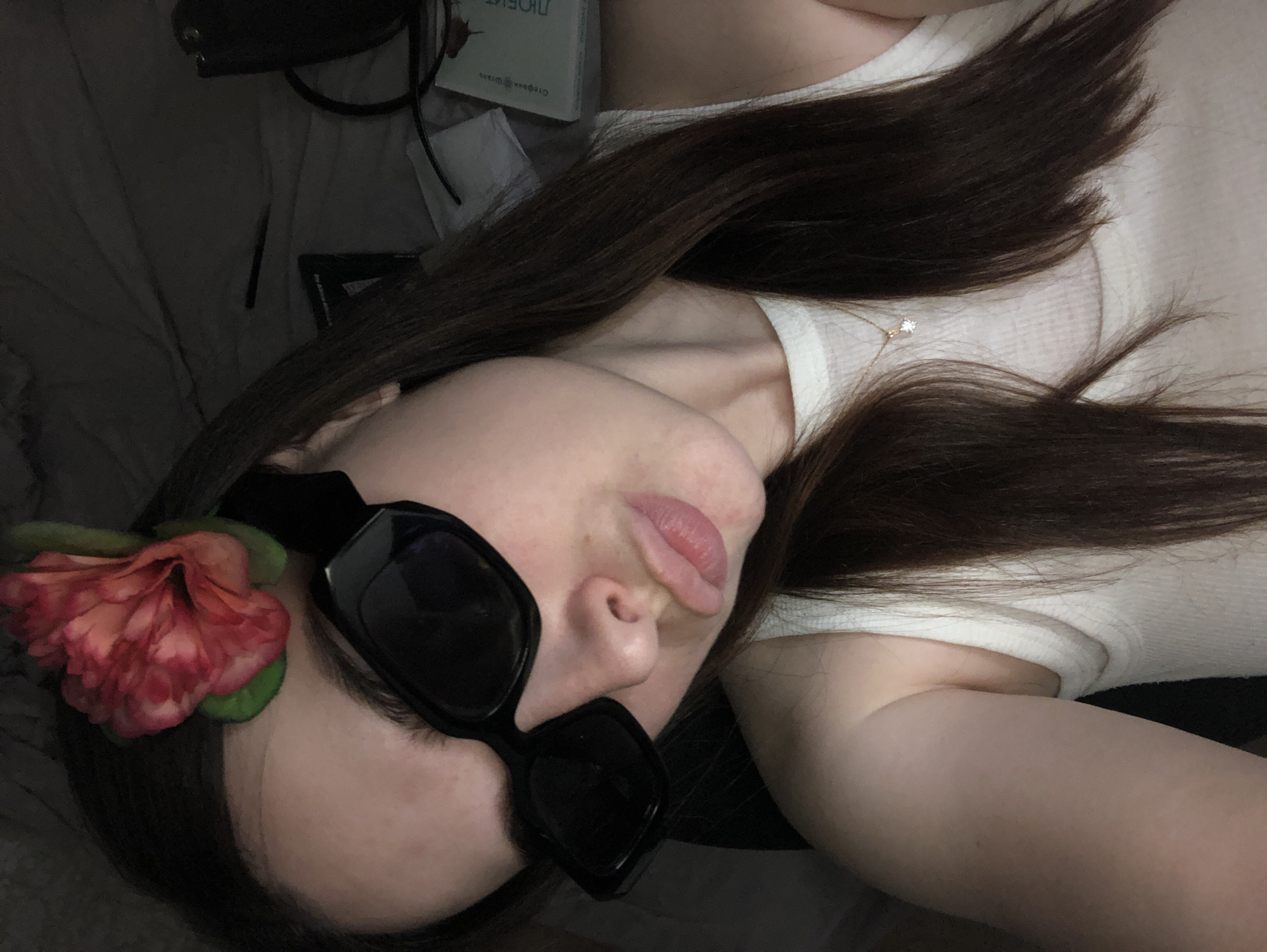 The image shows a woman lying down, likely on a bed. She is wearing a white top and dark sunglasses, with a flower attached to the side of the sunglasses. She has long, dark brown hair. A gold necklace is visible. A book and other miscellaneous items are in the background, in a dark, cluttered area. There is no evidence to suggest that the location is in the city of Noviy, Russia.
