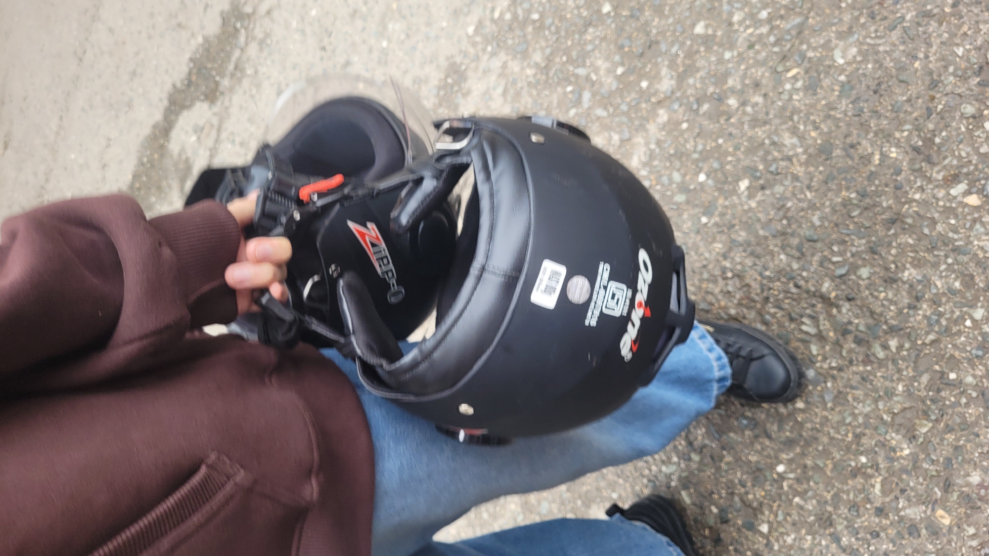 A person is holding a black motorcycle helmet in their lap, resting on their blue jeans. The helmet is open, revealing the black padding and straps inside. A clear visor is attached to the front. The person is wearing a brown hooded sweatshirt. The scene appears to be outdoors, with a rough, grey, pebbly ground surface visible. The lighting suggests it is daytime. There is visible text on the helmet, including a red and white logo that says "ZIPPER-O", a white sticker with text that is difficult to read but appears to be a certification mark and numbers like "RALS 1020", and a logo that says "Ozone" in white with a red accent. The setting is likely a street or parking area in Vashisht, India, given the ground texture. No specific event is clearly depicted, beyond the person preparing to use or having just used the helmet.
