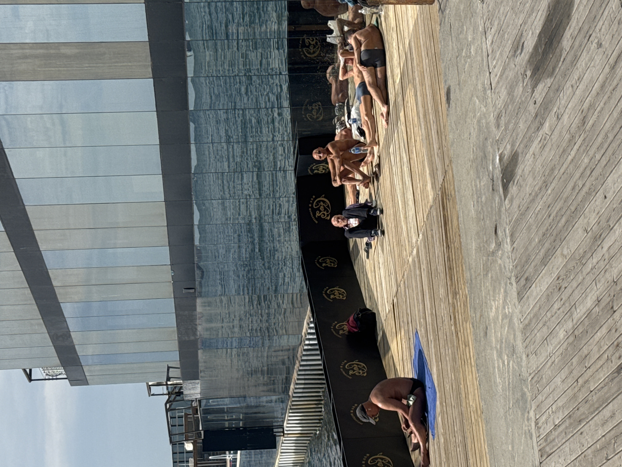 The image depicts a boardwalk scene in Vladivostok, Russia. A glass building reflects the sea. Along the boardwalk, several men in swimwear are gathered. One man in a suit is present. Another man stands on a blue towel. The boardwalk is made of wooden planks.
