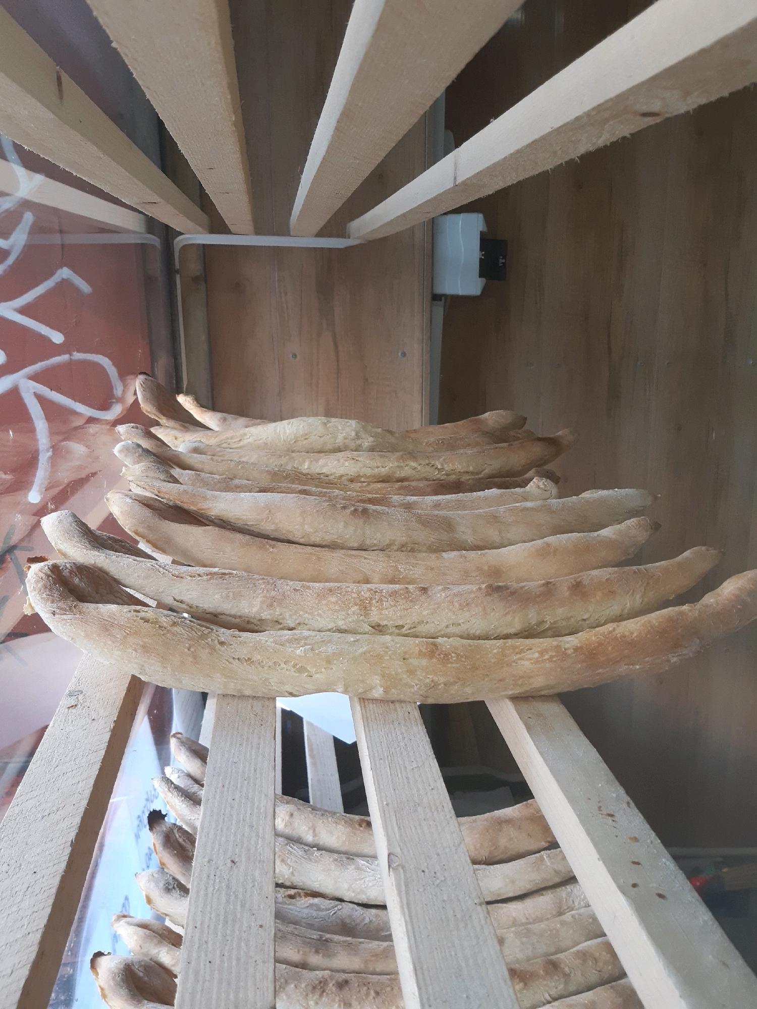 The image shows a close-up, eye-level, vertical shot of several loaves of bread stacked on wooden shelves. The bread appears to be of a long, slender baguette-like shape with a slightly golden-brown, textured crust. The shelves are made of raw, unfinished light-colored wood and are constructed in a way that creates a tiered effect, with the bread resting on the horizontal planks. In the background, there is a wooden wall and a portion of a red surface with white graffiti. To the right on the wooden wall, a white electrical box is visible. The lighting suggests an indoor setting, possibly a bakery or a market stall, with natural light coming from the left. The overall impression is one of rustic presentation of baked goods. The specific location in the city of Tbilisi, Georgia, cannot be determined from the visual information provided in the image.