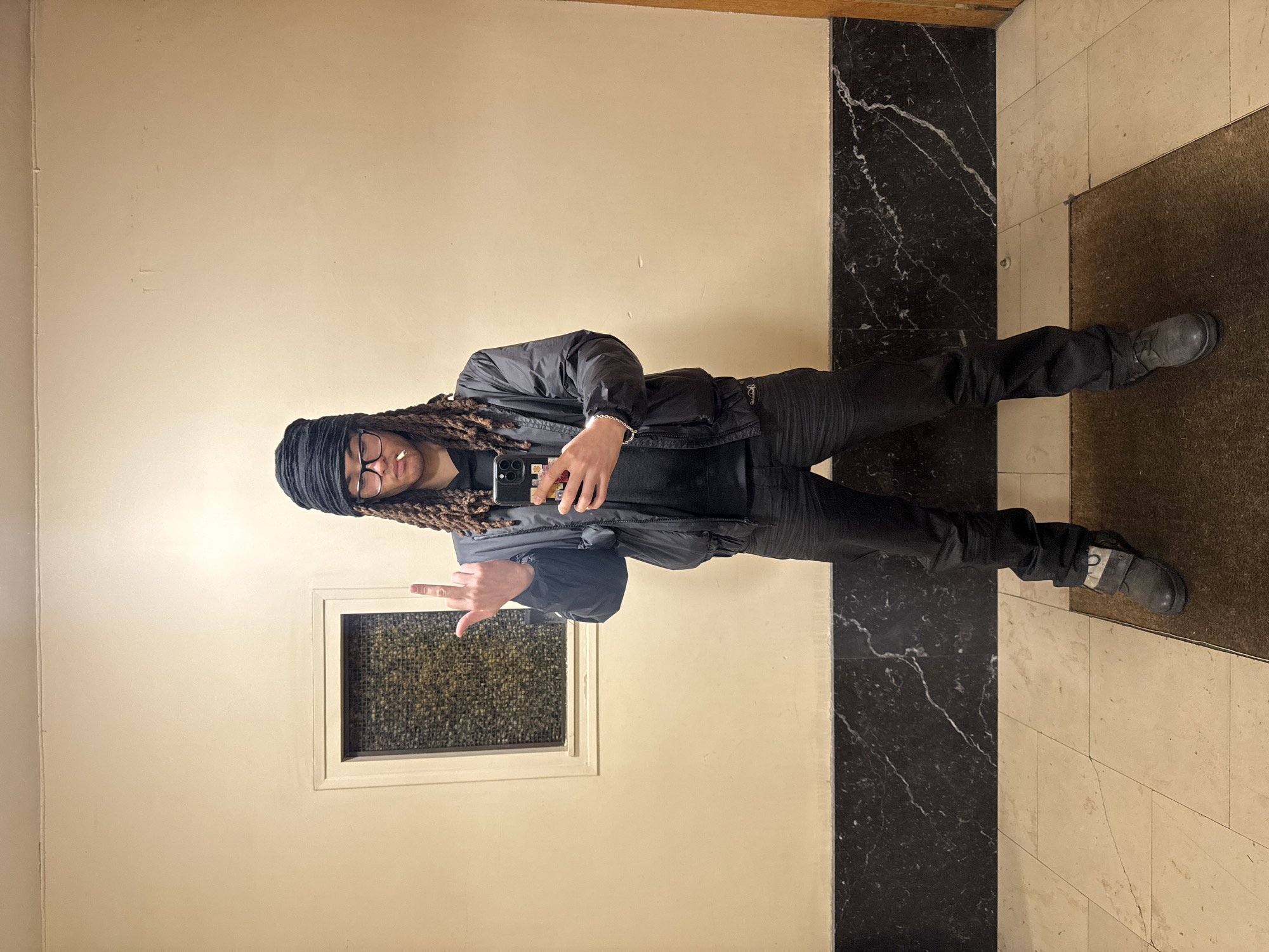 An individual stands centered in an interior hallway, consistent with the architectural style of residential or commercial buildings found in Paris, France. The subject, a person with dreadlocks, wears a black head covering, dark-framed glasses, a black puffer jacket over a black hooded sweatshirt, black pants, and dark boots with a strap detail. A lit cigarette is present in their mouth. The individual holds a black smartphone with a multi-colored sticker-covered case in their right hand, angled as if capturing a mirror selfie. Their left hand is raised, with the middle finger extended upwards. The background features light beige walls above a dark, veined marble wainscoting. To the left, a rectangular opening with textured, opaque glass is set into the wall. The floor is composed of light-colored square tiles, and a dark brown mat occupies the lower right foreground. A wooden door frame is partially visible on the extreme right.