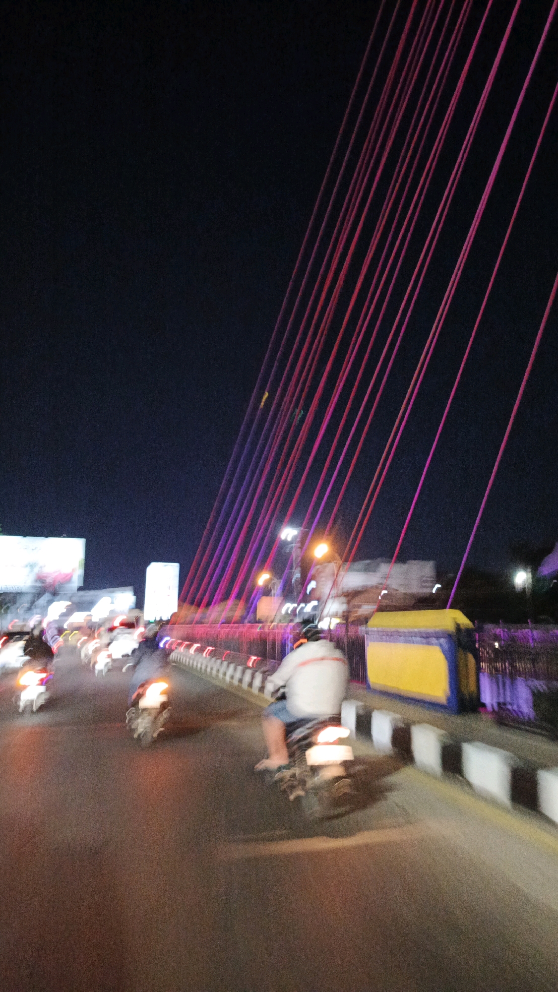 The image shows a view from a motorcycle on a bridge at night in Surakarta, Indonesia. The bridge is illuminated with pink and purple lights that are strung along its cables. Several motorcycles are visible on the bridge, with one rider in the foreground. In the background, there are buildings and streetlights. The overall scene is one of movement and nighttime illumination.