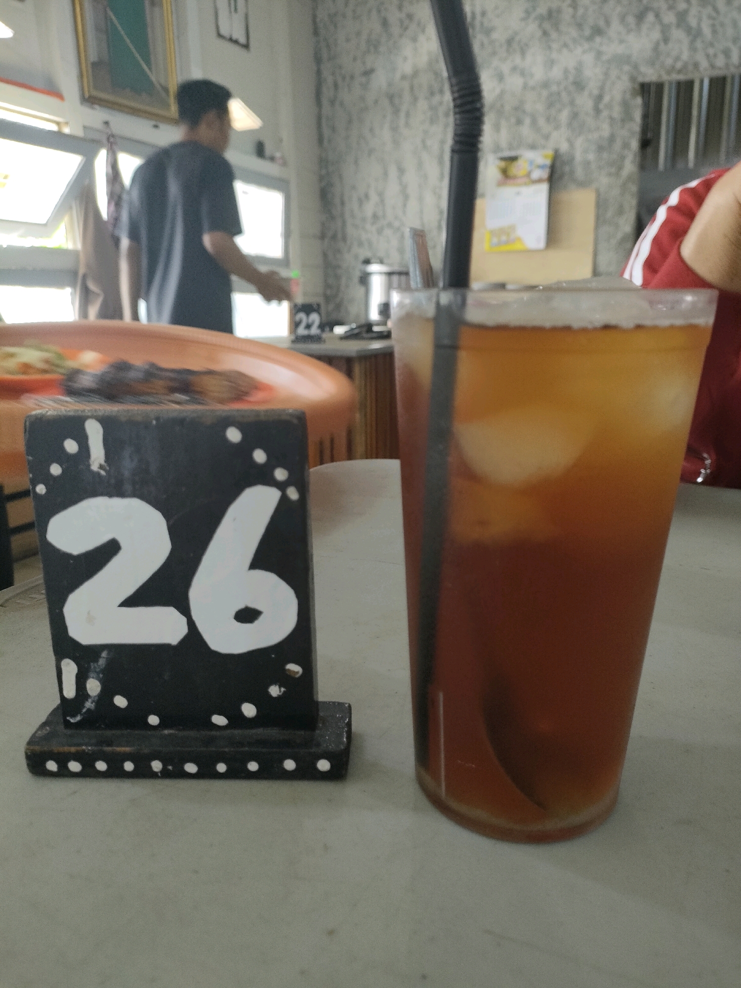 In an indoor setting in Banjarmasin, Indonesia, a table is set with food and drinks. In the foreground, a black table marker with white numbers "26" is prominently displayed. To the right, a tall glass of iced tea with a black straw stands. In the background, a person in a dark t-shirt is visible near a window, and another person wearing a red and white striped shirt is partially visible. The environment appears to be a casual dining area, possibly a restaurant or a cafe, with a textured wall in the background and a framed picture hanging on it. The lighting suggests it is daytime.