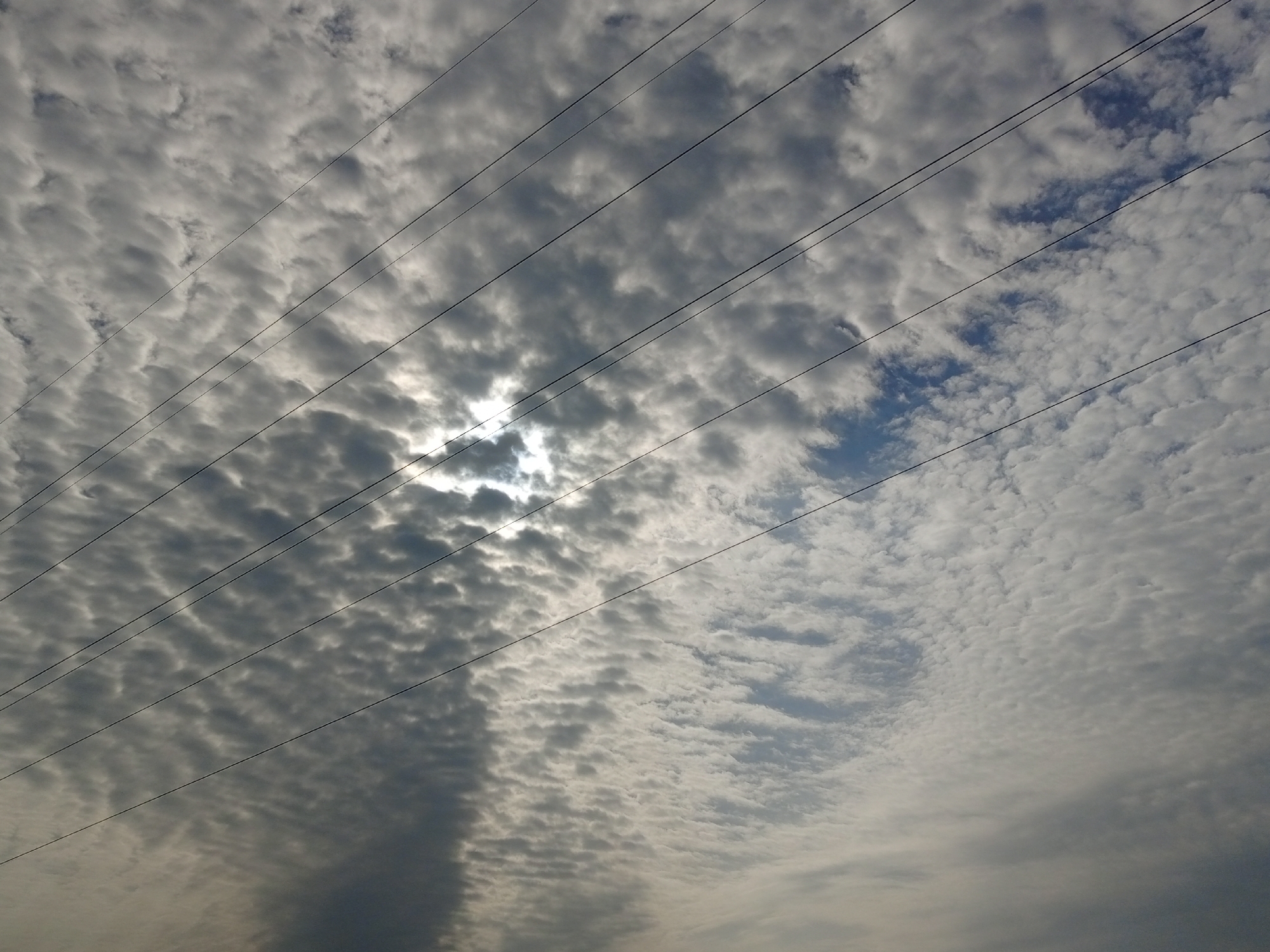 The image depicts a cloudy sky with several overhead power lines stretching diagonally across the frame. The clouds are varied, with some appearing dense and dark, while others are lighter and more wispy, allowing patches of blue sky to peek through. The overall lighting suggests it might be overcast or during a time of day when the sun is not directly overhead. The power lines are thin and black, creating a linear pattern against the textured backdrop of the clouds. The scene appears to be an outdoor environment, potentially urban or semi-urban, given the presence of the power lines. No people or specific identifiable landmarks are visible. The location is identified as Rukuba Road, Nigeria.