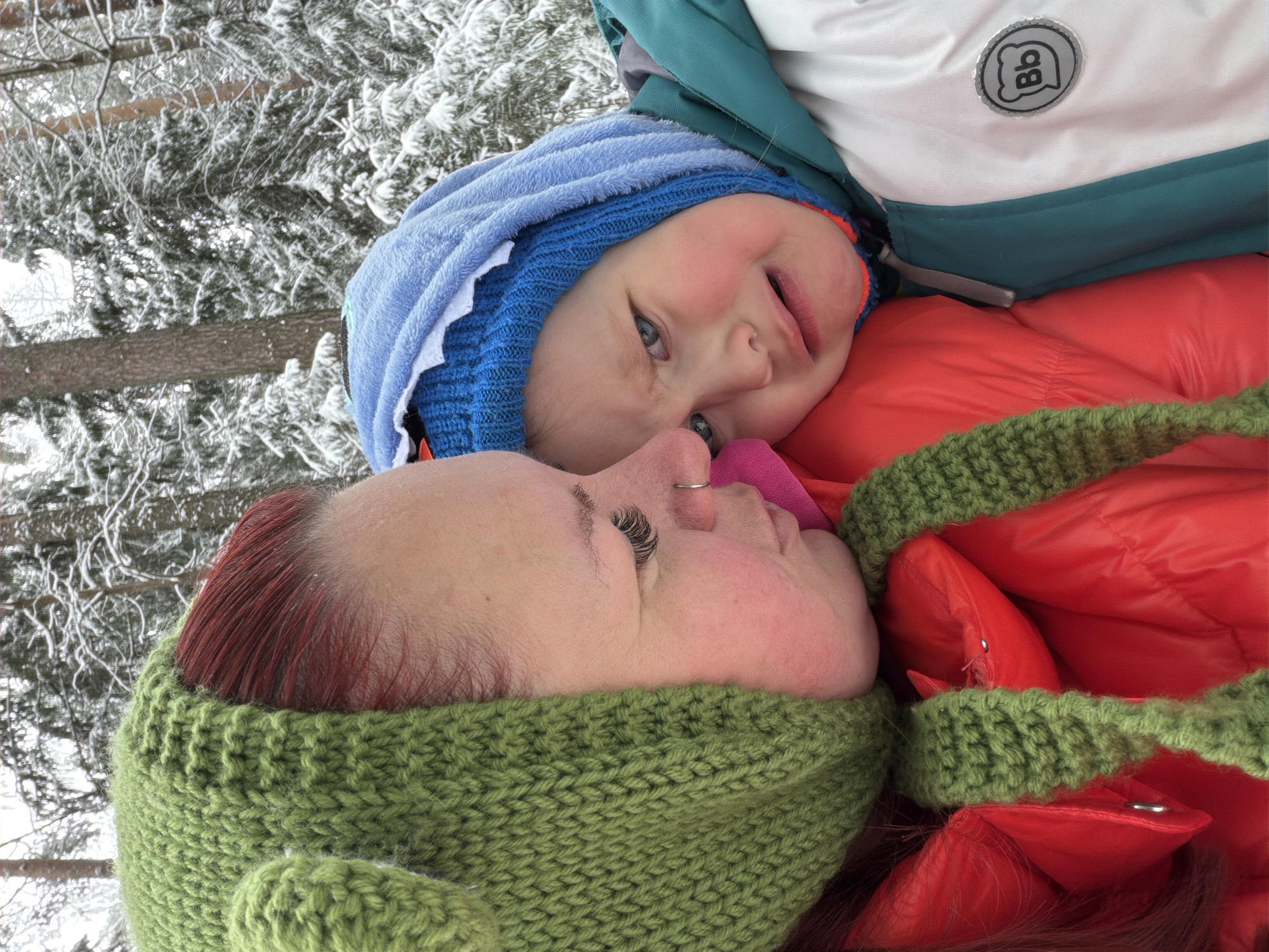 A photograph taken in what appears to be a snowy outdoor environment in the city of Chaikovsky, Russia, depicts a red-haired woman wearing a green knitted hat lying with a small child on top of her. The child is wearing a blue hat and is laying on top of a red winter jacket with green trim. Snow-covered trees are visible in the background.