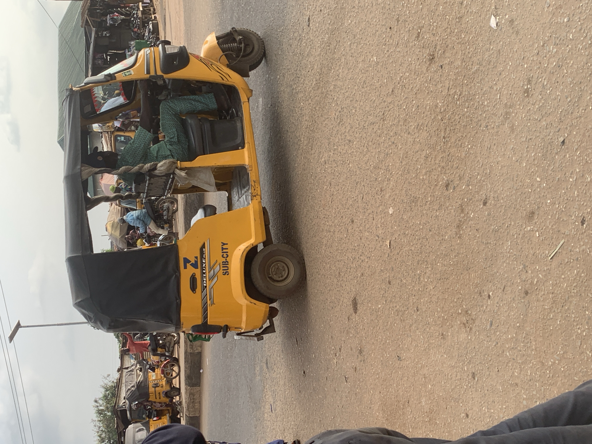 In Shagamu, Nigeria, a yellow auto-rickshaw, a common mode of transport known as a "Keke napep," is parked on a dusty road. The auto-rickshaw has a black fabric roof and "DELUXE PLUS SUB-CITY" written on its side. A driver, dressed in a green patterned outfit and a black cap, is seated behind the steering wheel, with a braided piece of fabric hanging from the back of the seat. In the background, other auto-rickshaws and motorcycles are visible, as well as market stalls with green roofing and various goods displayed. The sky is overcast. The scene depicts a typical day in a Nigerian city.