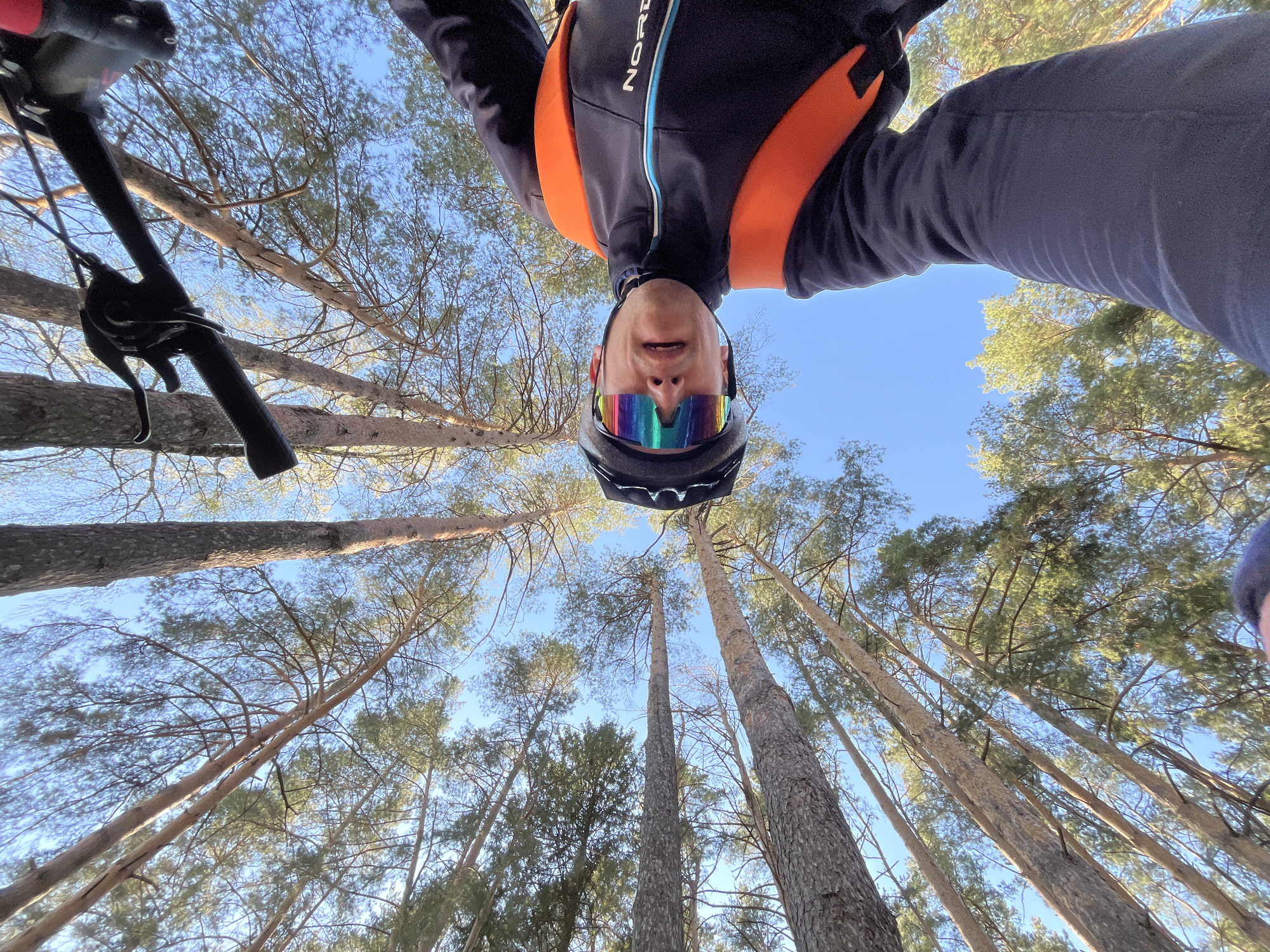 A low-angle, rotated photograph shows a person upside down in a forest, likely in the city of Chaikovsky, Russia. The individual is wearing a black bicycle helmet and iridescent sunglasses. They are also wearing a dark jacket with orange shoulder straps and the word "NORD" visible on the shoulder. The bike's handlebar is visible on the left side of the frame. Tall pine trees dominate the background, reaching towards a clear, blue sky.
